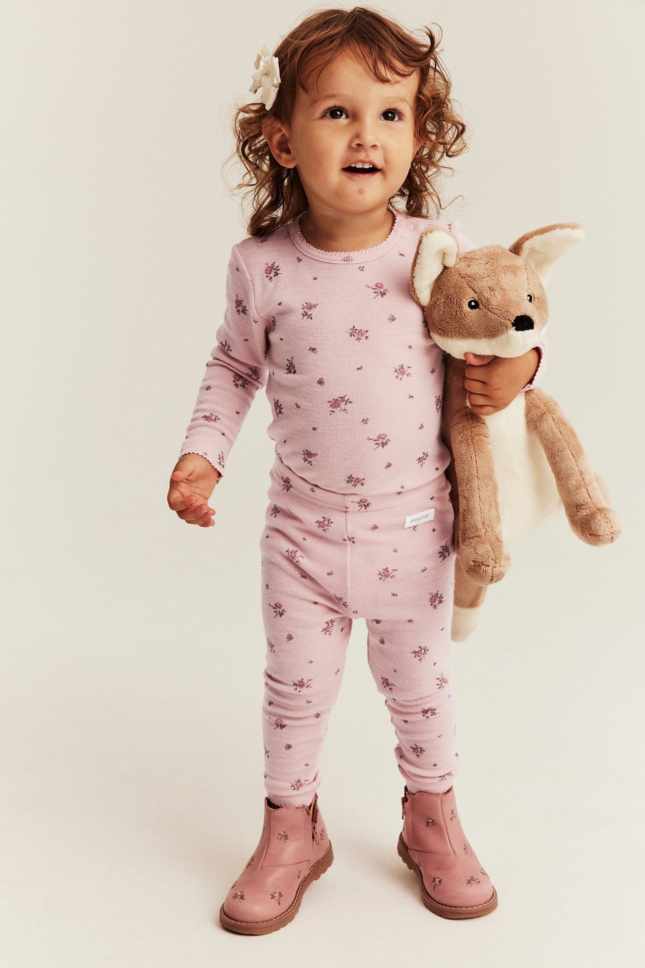 Front view of a kid wearing a pink floral long-sleeved pajama set, pink boots, and holding a brown stuffed animal.