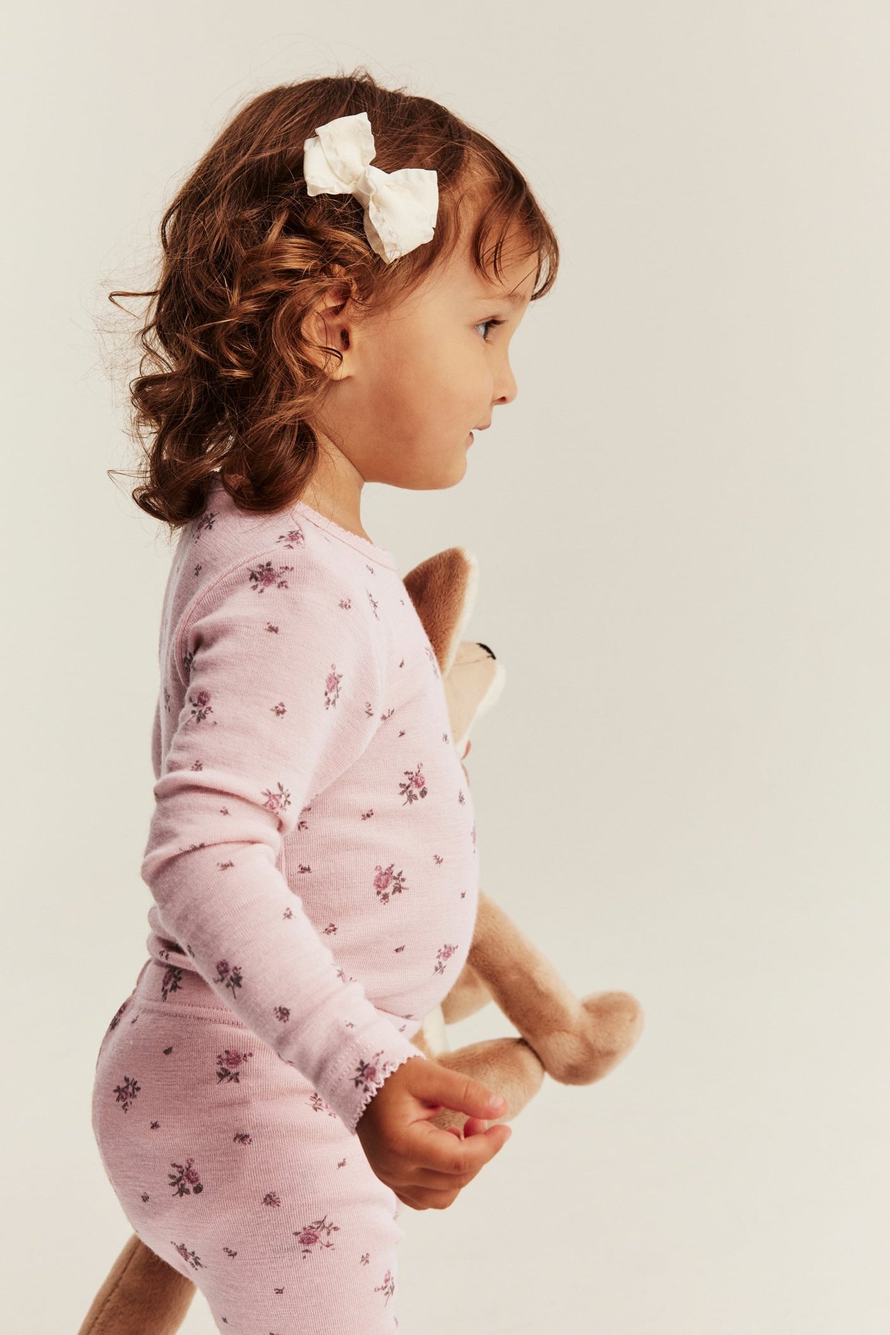Side view of a kid in a pink floral long-sleeved top and matching pants, holding a brown plush toy.