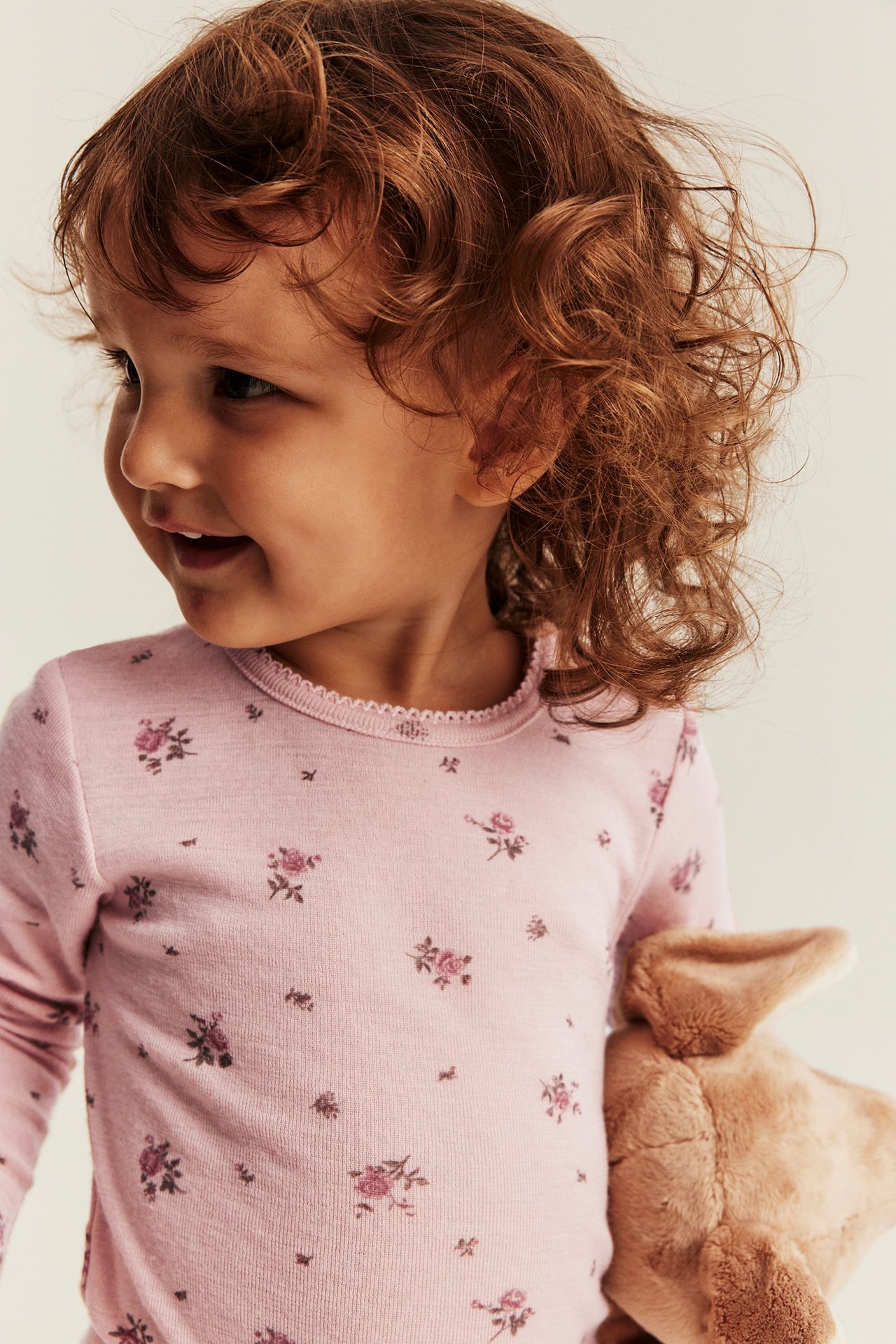 Close-up of a kid in a pink floral long-sleeved top with scalloped neckline, holding a brown plush toy.