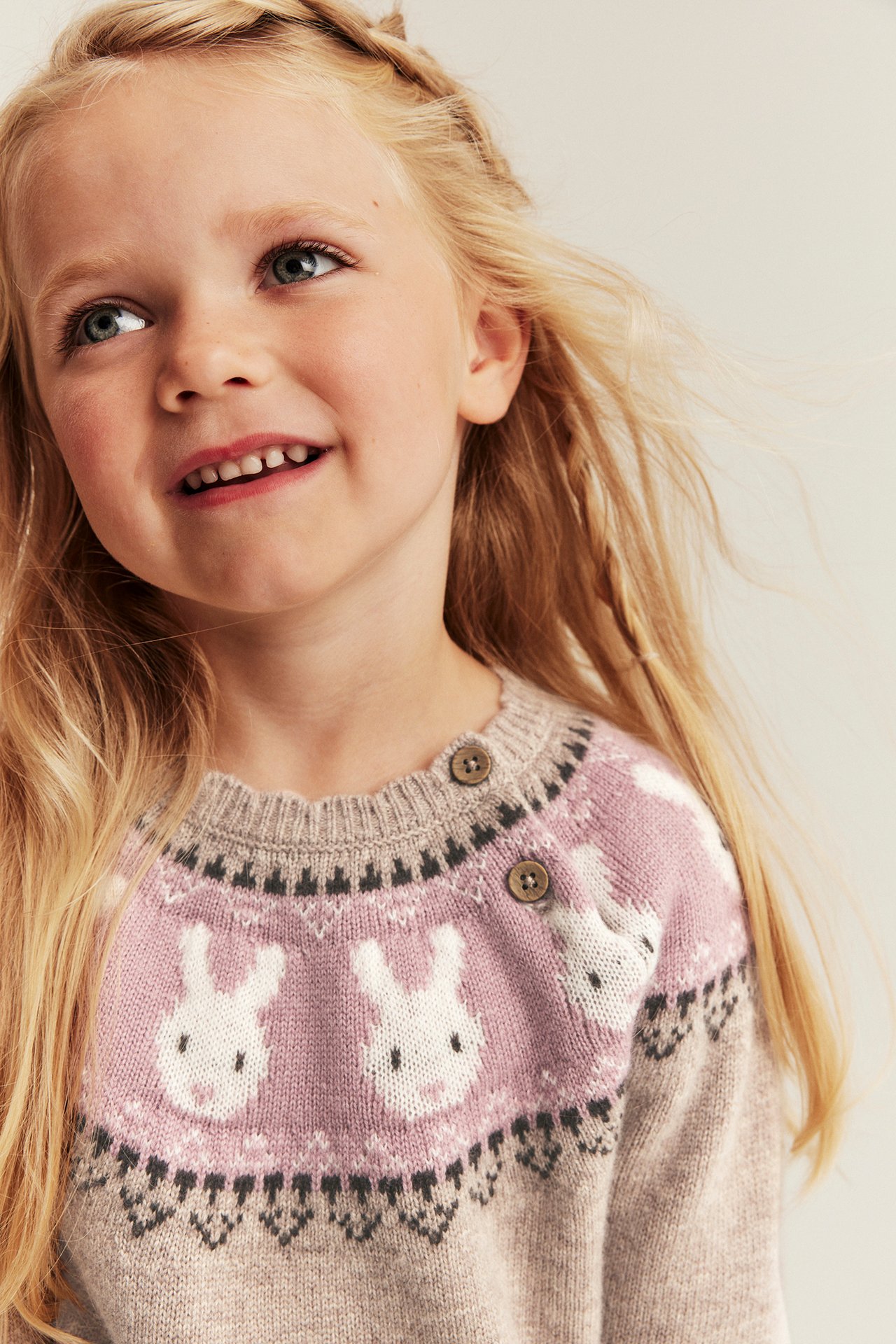 Nahaufnahme eines Kindes in einem rosa-beigen Fair Isle Hasenpullover mit Schulterknöpfen.