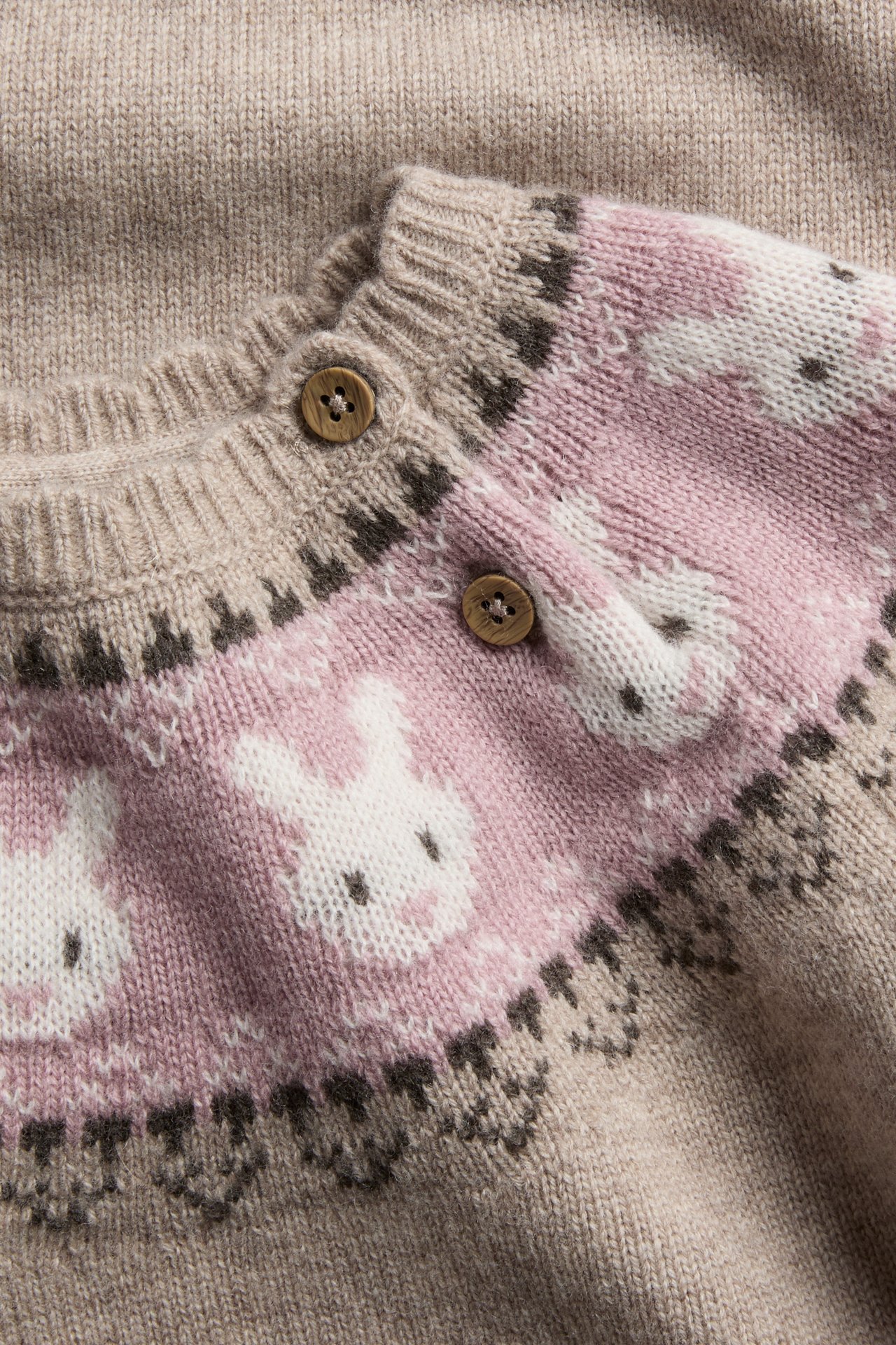 Nahaufnahme einer gestrickten Kinderstrickjacke mit beigem Grund, rosa Hasenmuster und braunen Knöpfen.