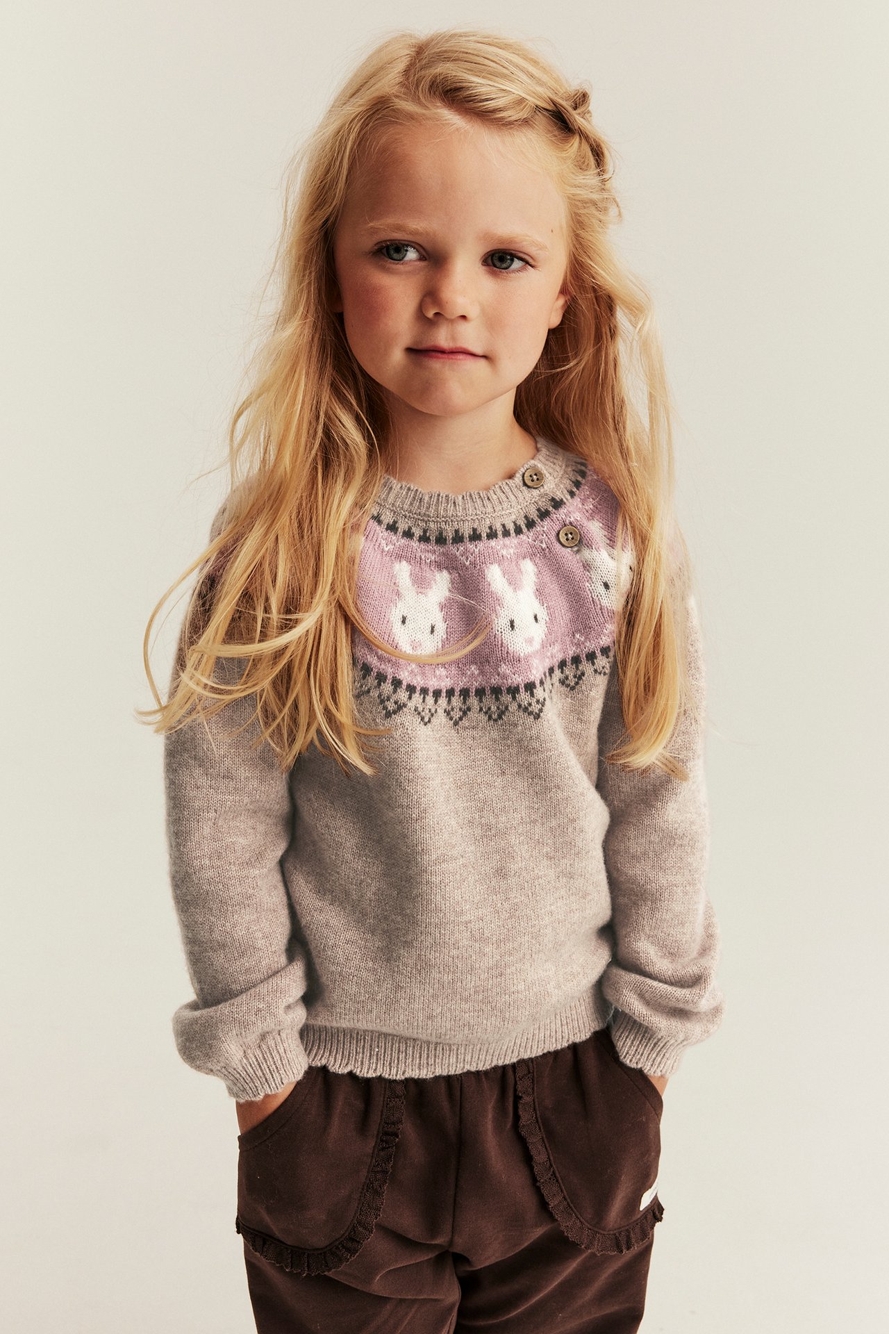 Vorderansicht eines Mädchens in beigem Strickpullover mit rosa Hasenmuster und brauner Cordhose.