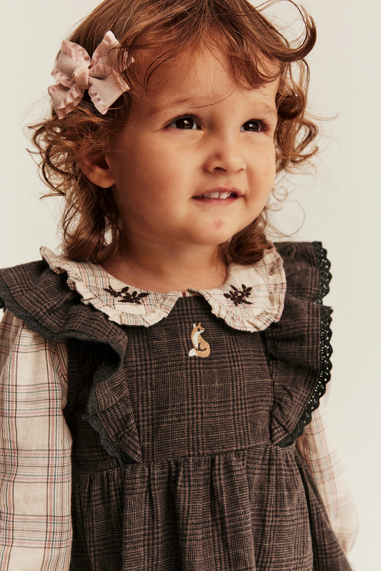 Kid in dark brown plaid overall dress with fox, light plaid shirt, pink bow.