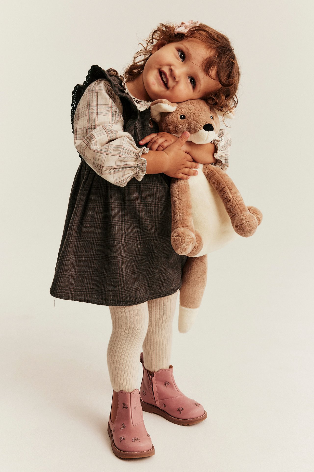 Kid wears plaid pinafore dress over checkered blouse, ribbed tights, and pink embroidered ankle boots.
