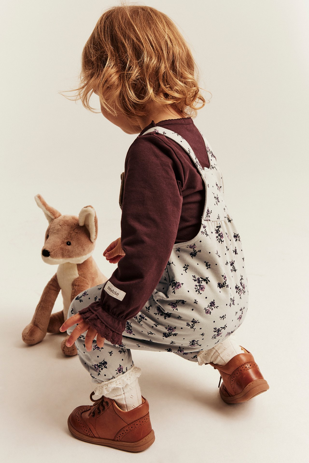 Back view, kid in light blue floral overall over a dark red long-sleeve top, white socks, brown shoes.