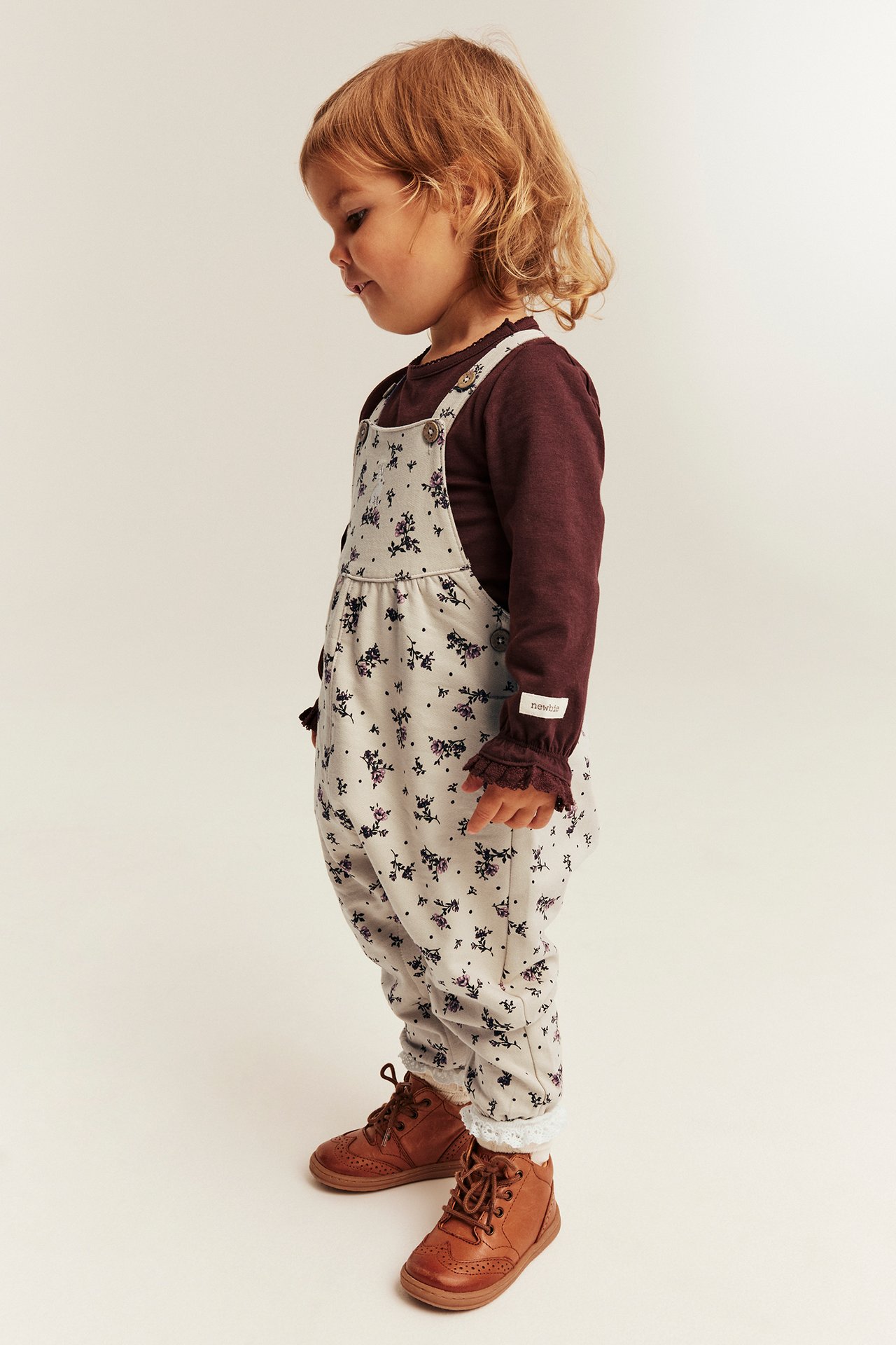 Side view: Kid in floral print overalls, burgundy long-sleeve top, and brown lace-up boots.