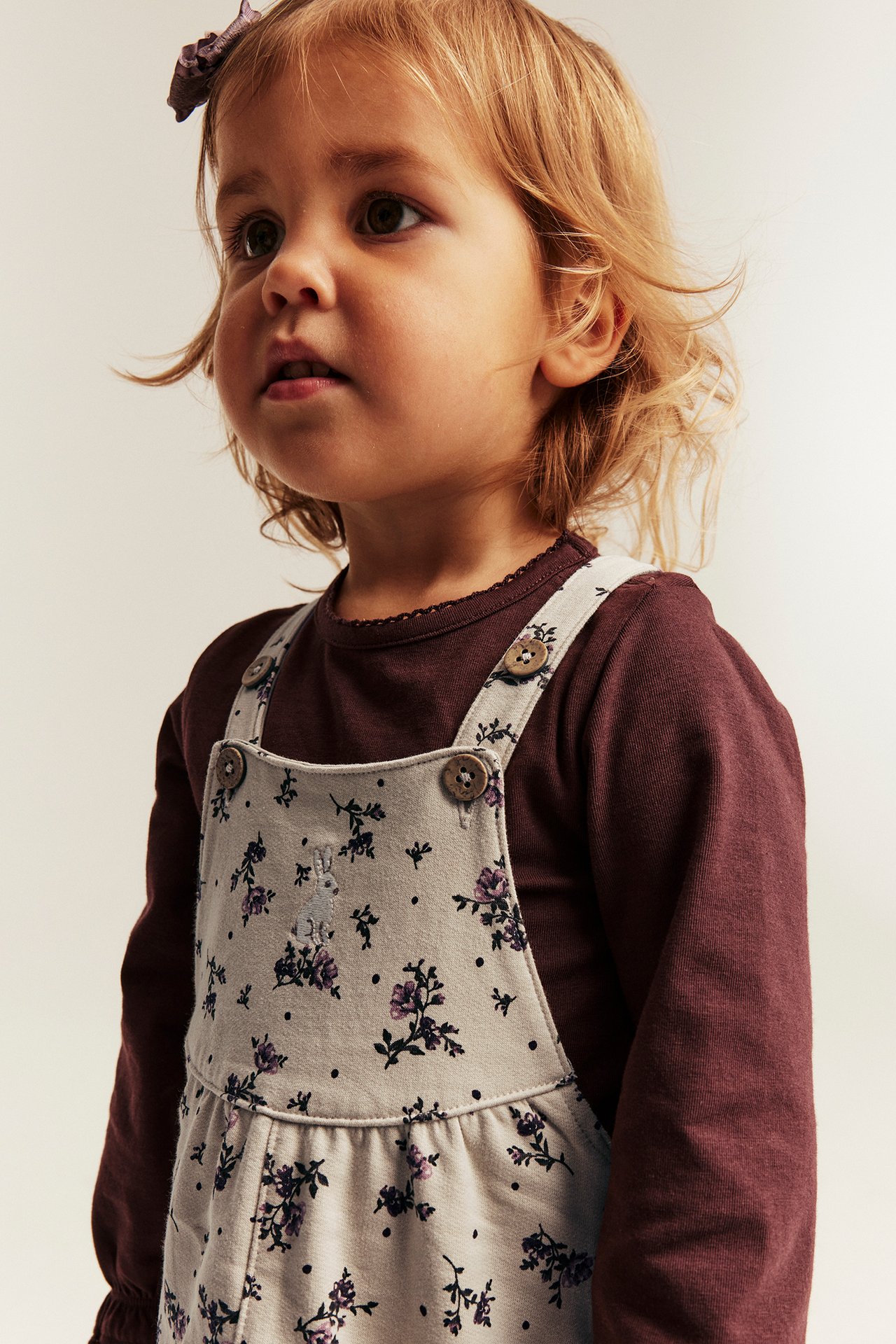 Front view of a kid in a floral beige overall with buttons over a dark brown long-sleeved top.