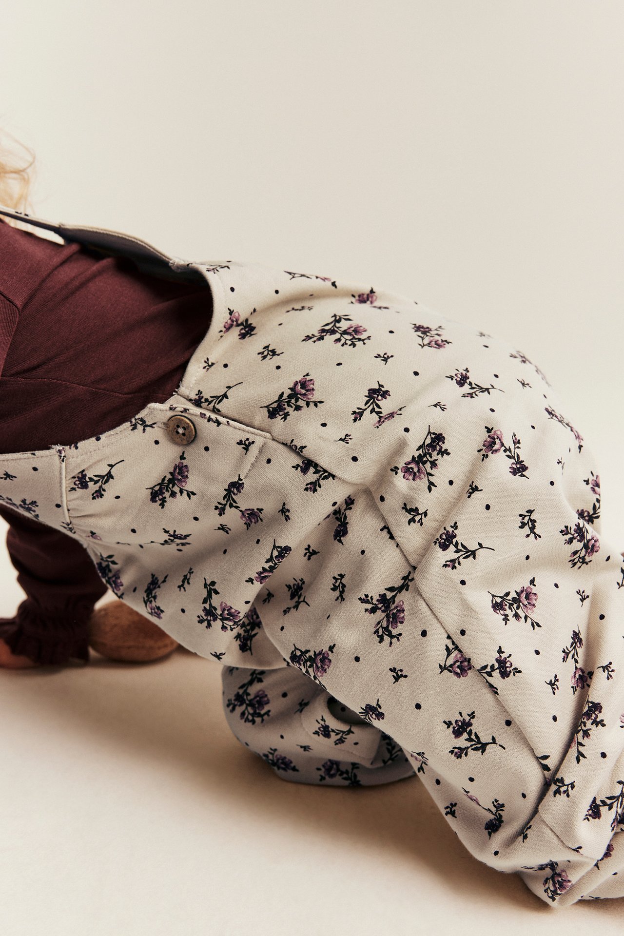 Close-up of a baby wearing a cream floral print overall over a dark red long-sleeved top.