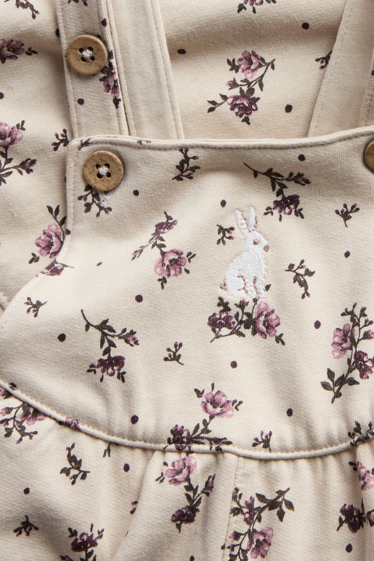 Close-up of a baby's beige floral print overall with a white bunny embroidery and wooden buttons.