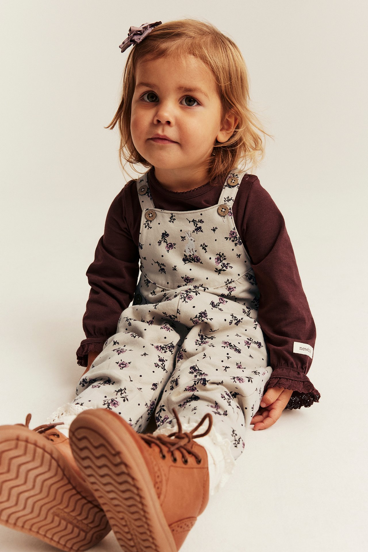 Front view of kid in light grey floral overall, dark brown long-sleeve top, brown lace-up boots, purple hair clip.