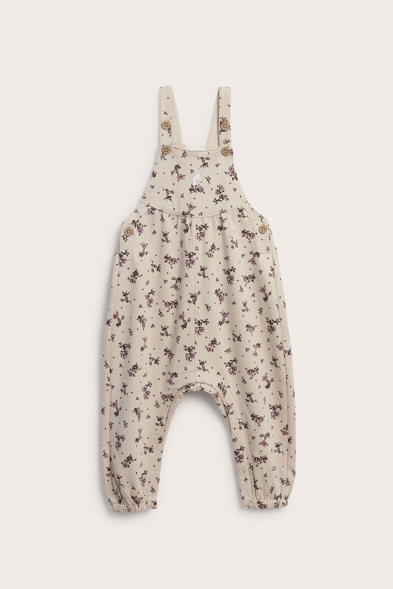 Front view of a baby's beige floral print overall with a white rabbit logo and button straps.