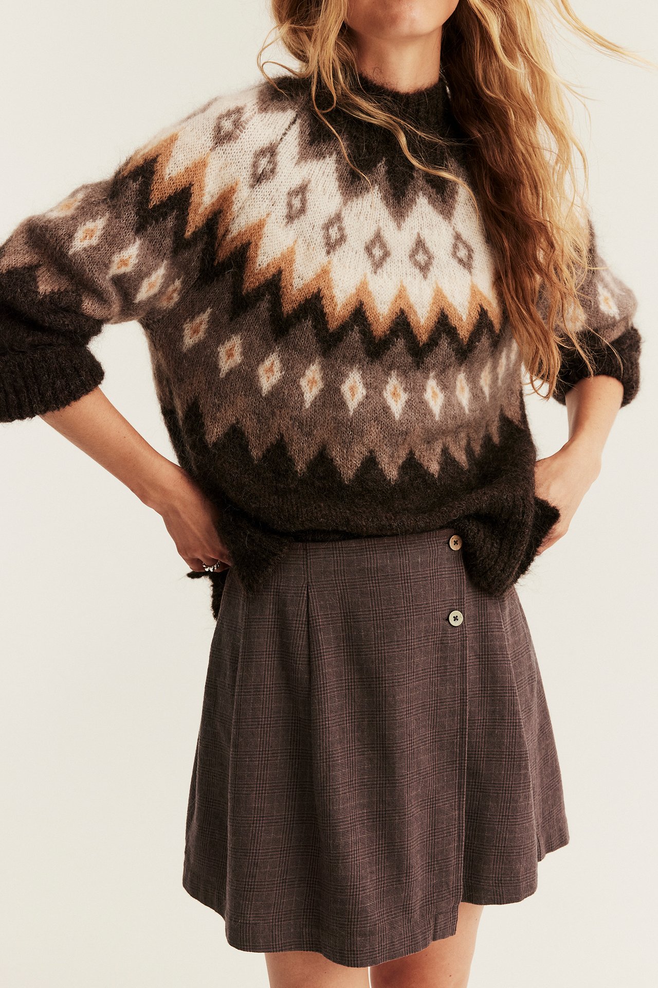Frontansicht Frau mit braunem Fair Isle-Pullover über dunklem Karo-A-Linienrock mit Knöpfen.