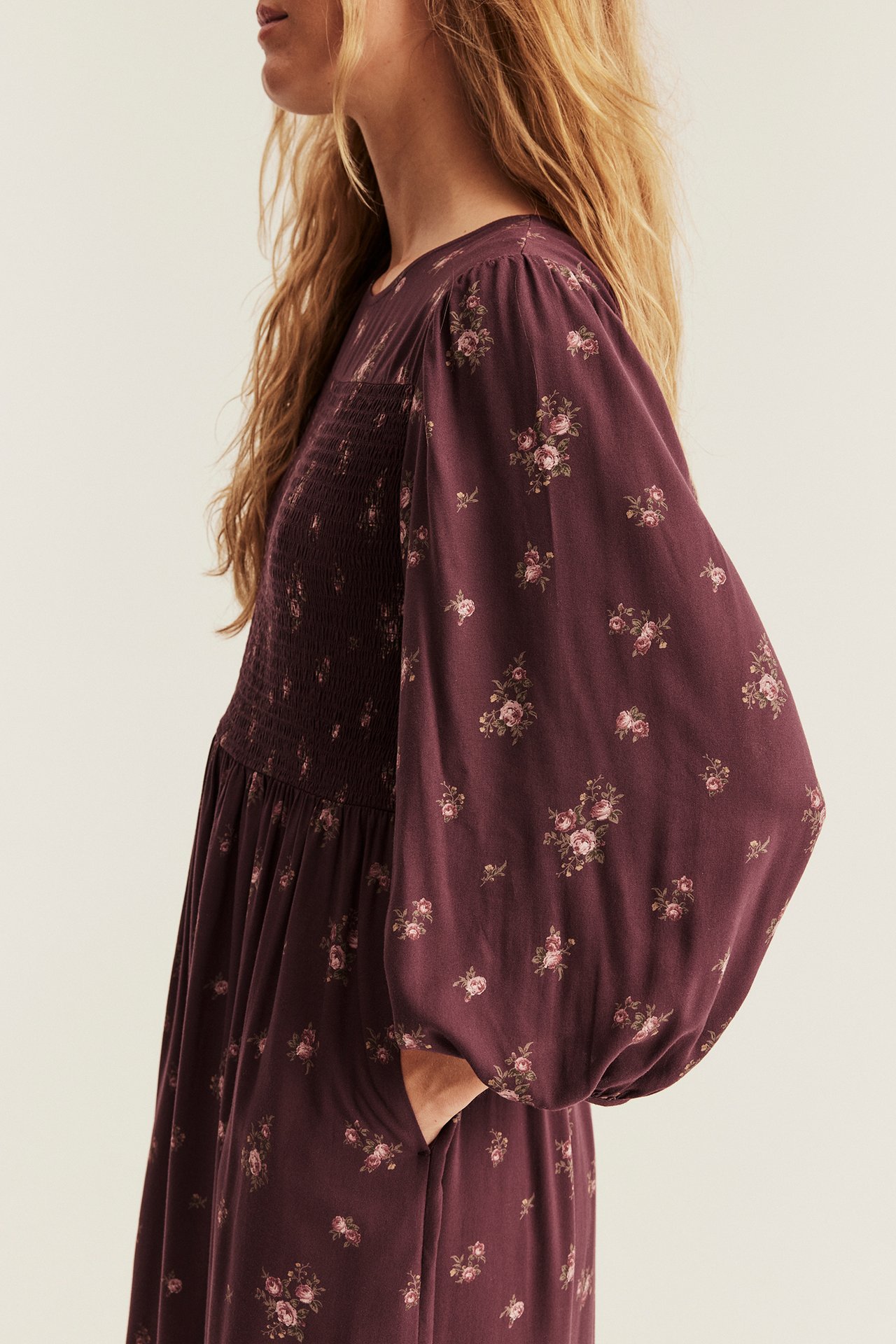 Side view, woman wearing a dark purple floral smocked long-sleeved dress with pockets.