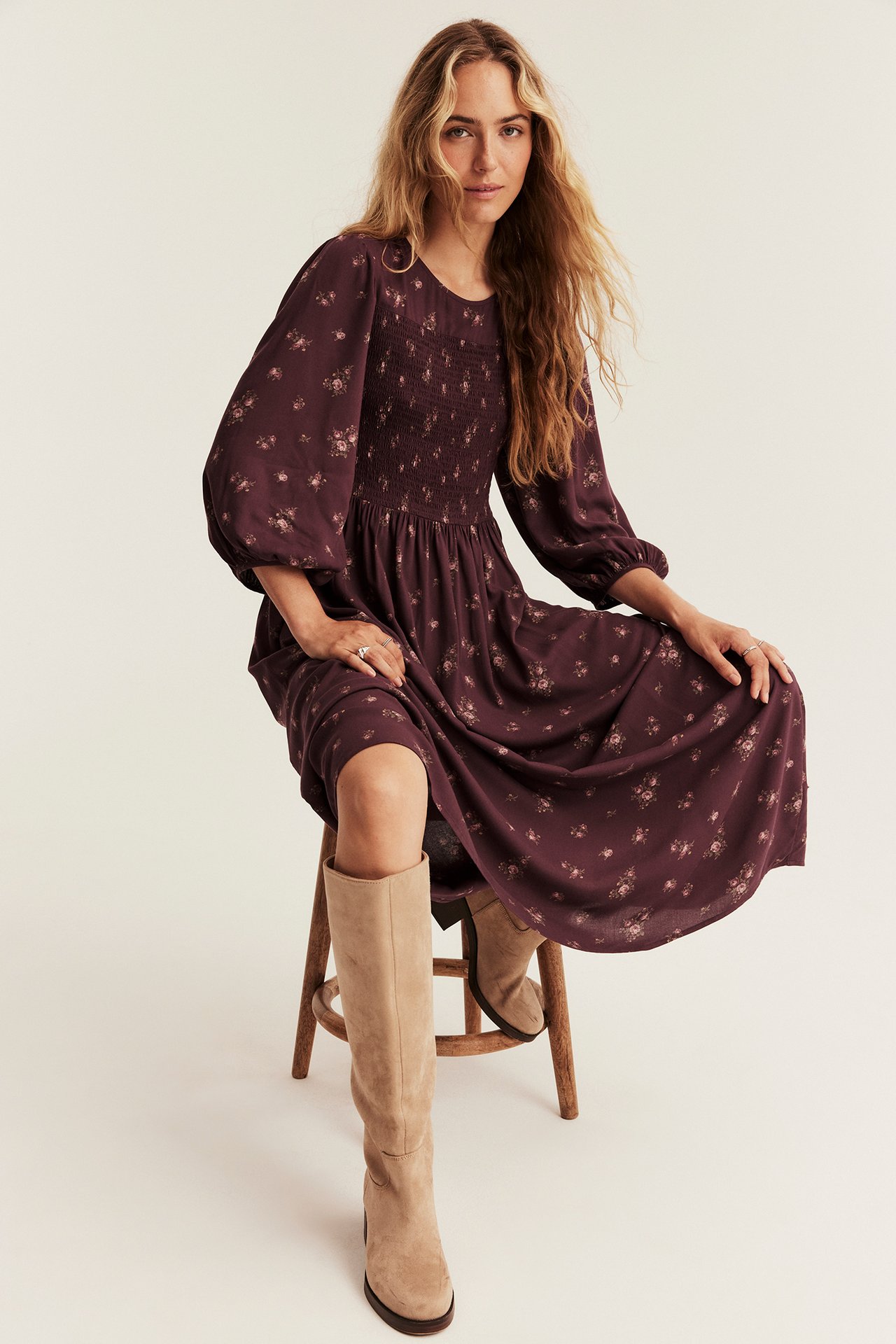 Front view of a woman wearing a dark purple floral smocked midi dress and beige suede knee-high boots.