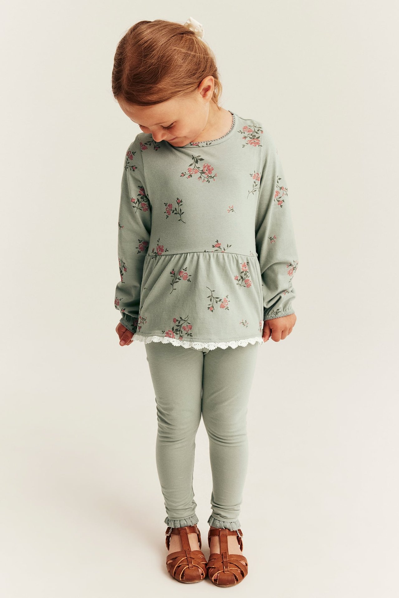 Vorderansicht: Ein Kind trägt ein grünes geblümtes Langarm-Peplum-Oberteil mit Spitzenrand, grüne Leggings und braune Sandalen.