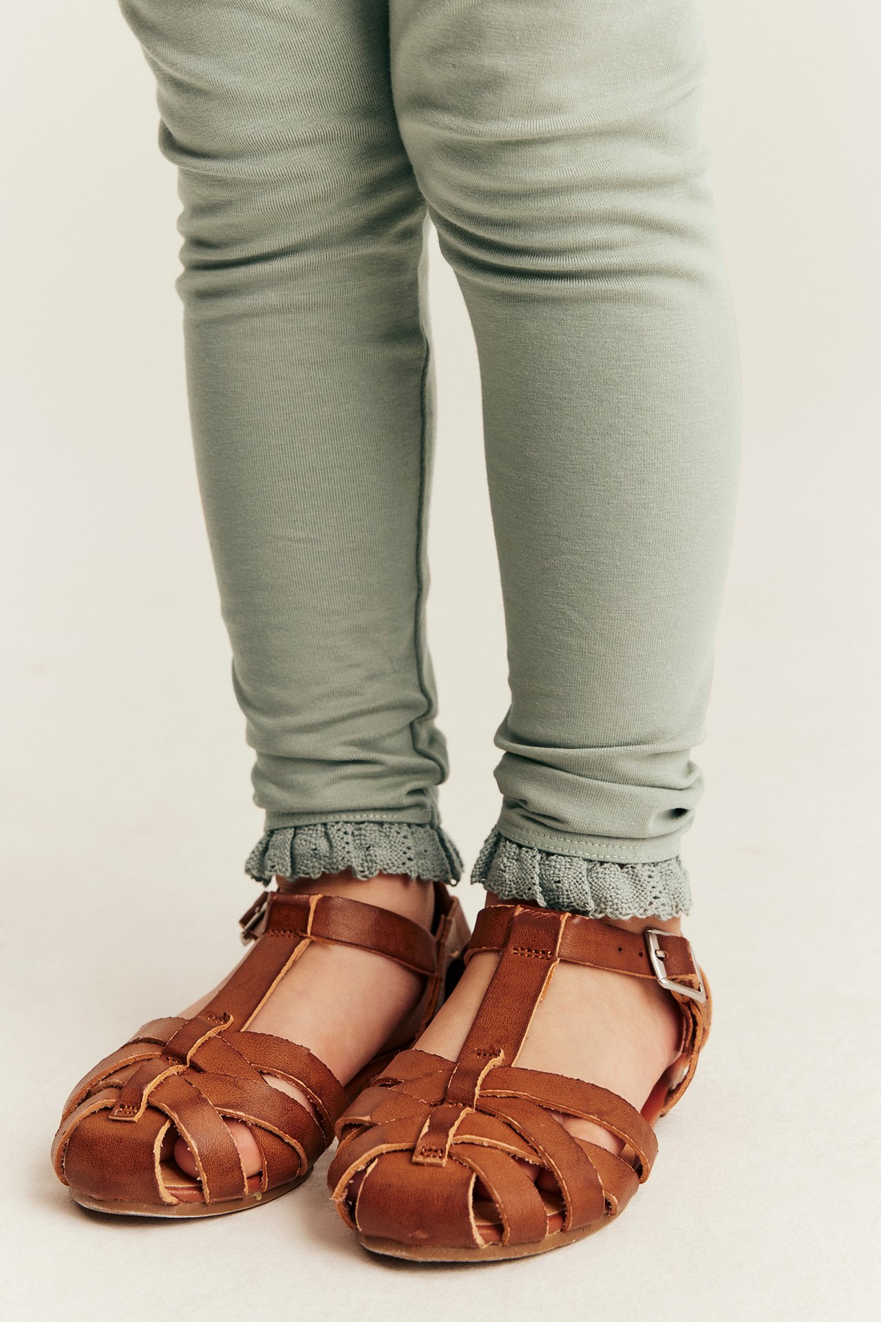 Close-up front view of a kid wearing light green leggings with frilly grey-blue lace trim and brown leather sandals.