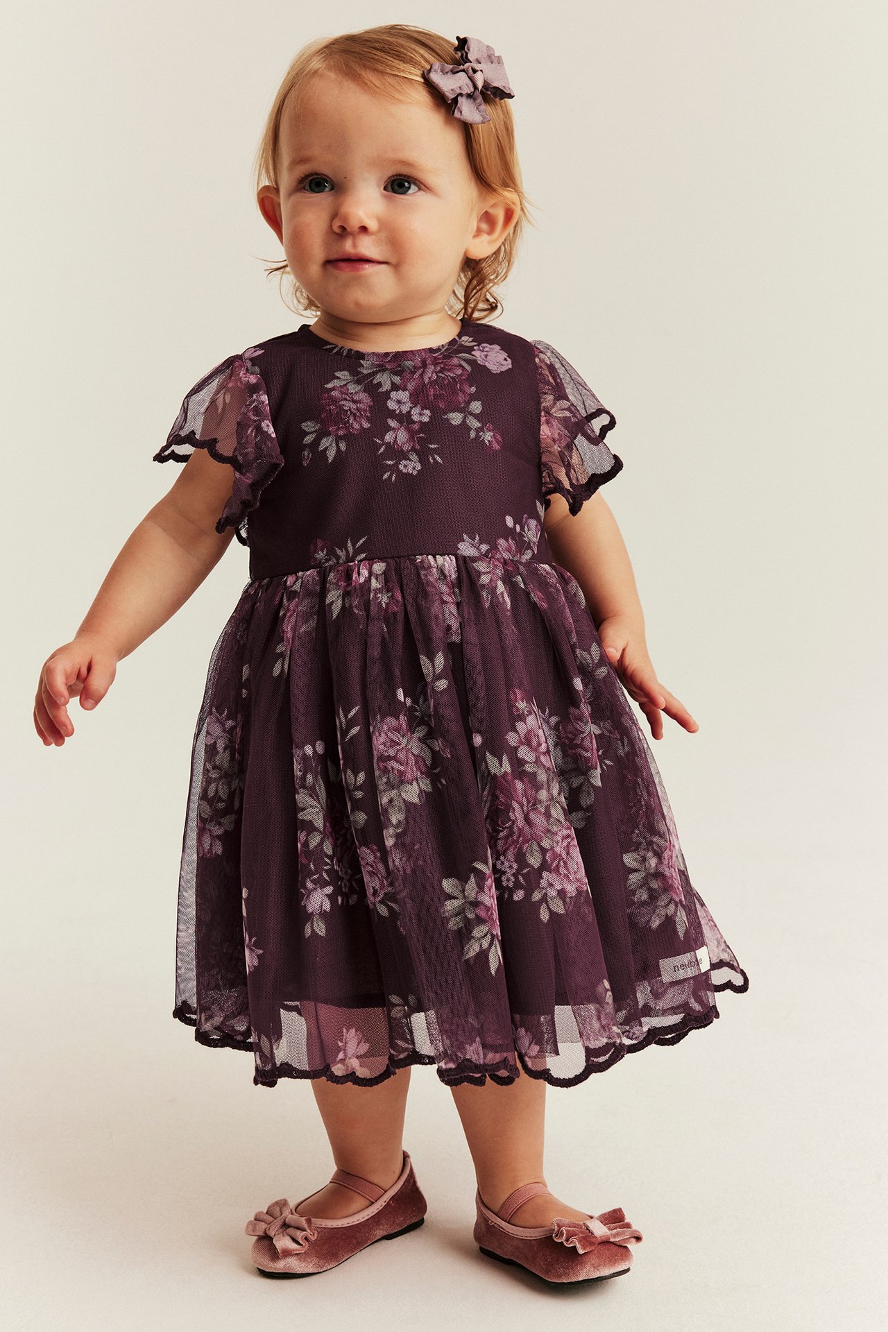 Vorderansicht eines Babys in einem dunkelvioletten Blumenkleid mit transparenten Ärmeln und rosa Samtballerinas.