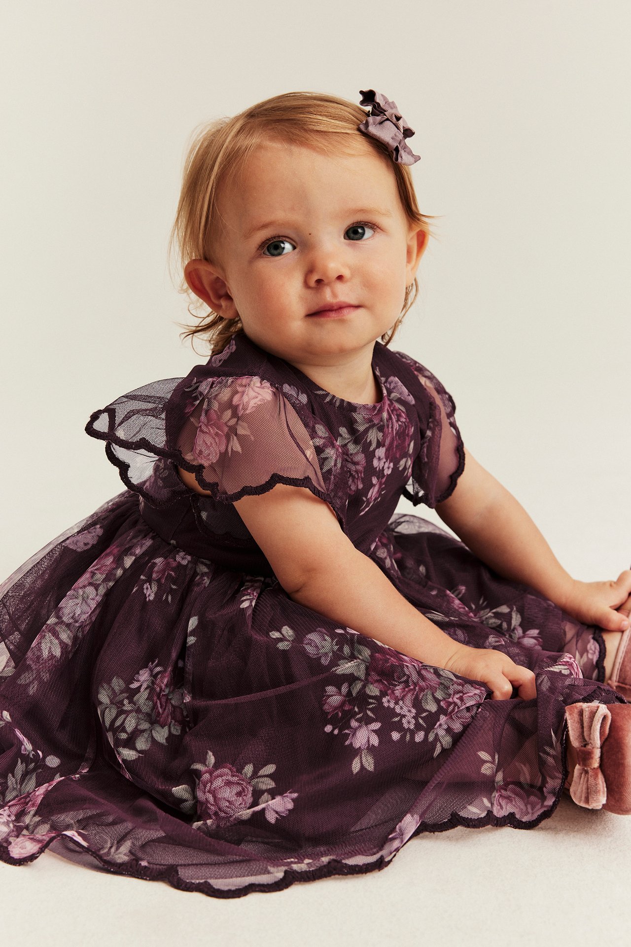 Vorderansicht: Baby in dunkelviolettem Blumen-Tüllkleid, passender Haarspange und rosa Samtschuhen.