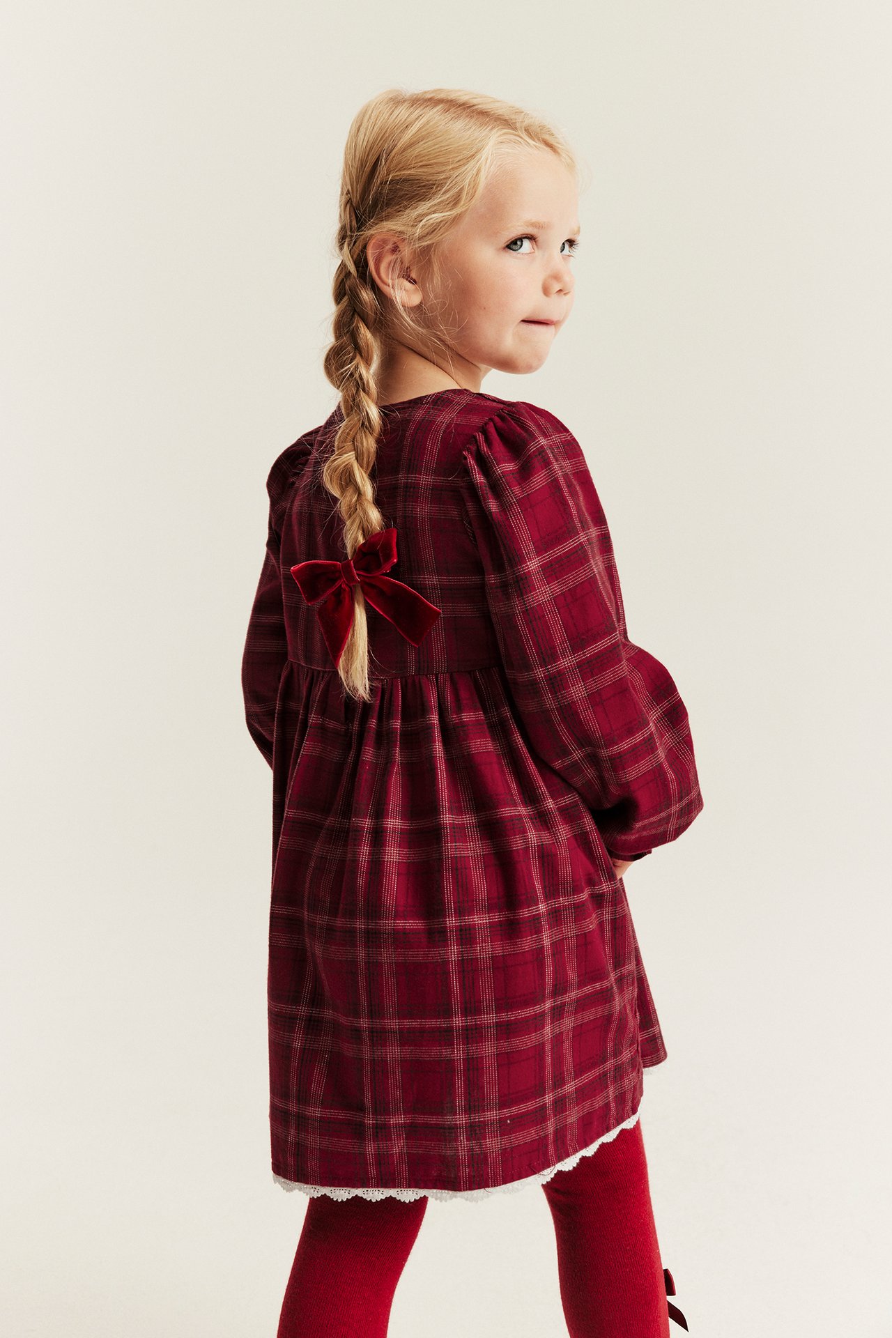 Side view of a kid wearing a red plaid long-sleeved dress with a red velvet bow and red tights.