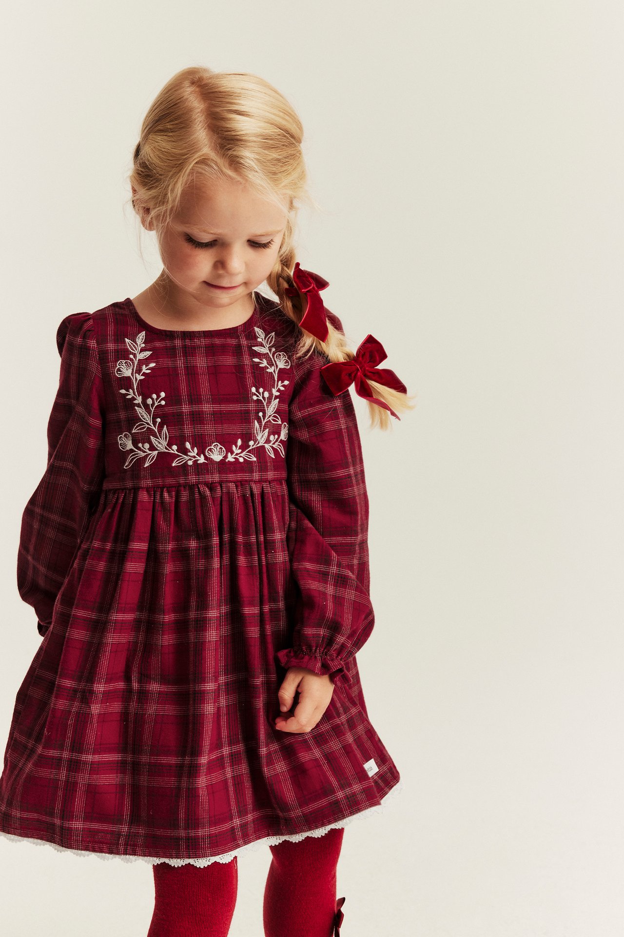 Kid's long-sleeved maroon plaid dress with embroidery, red tights, and hair bows.