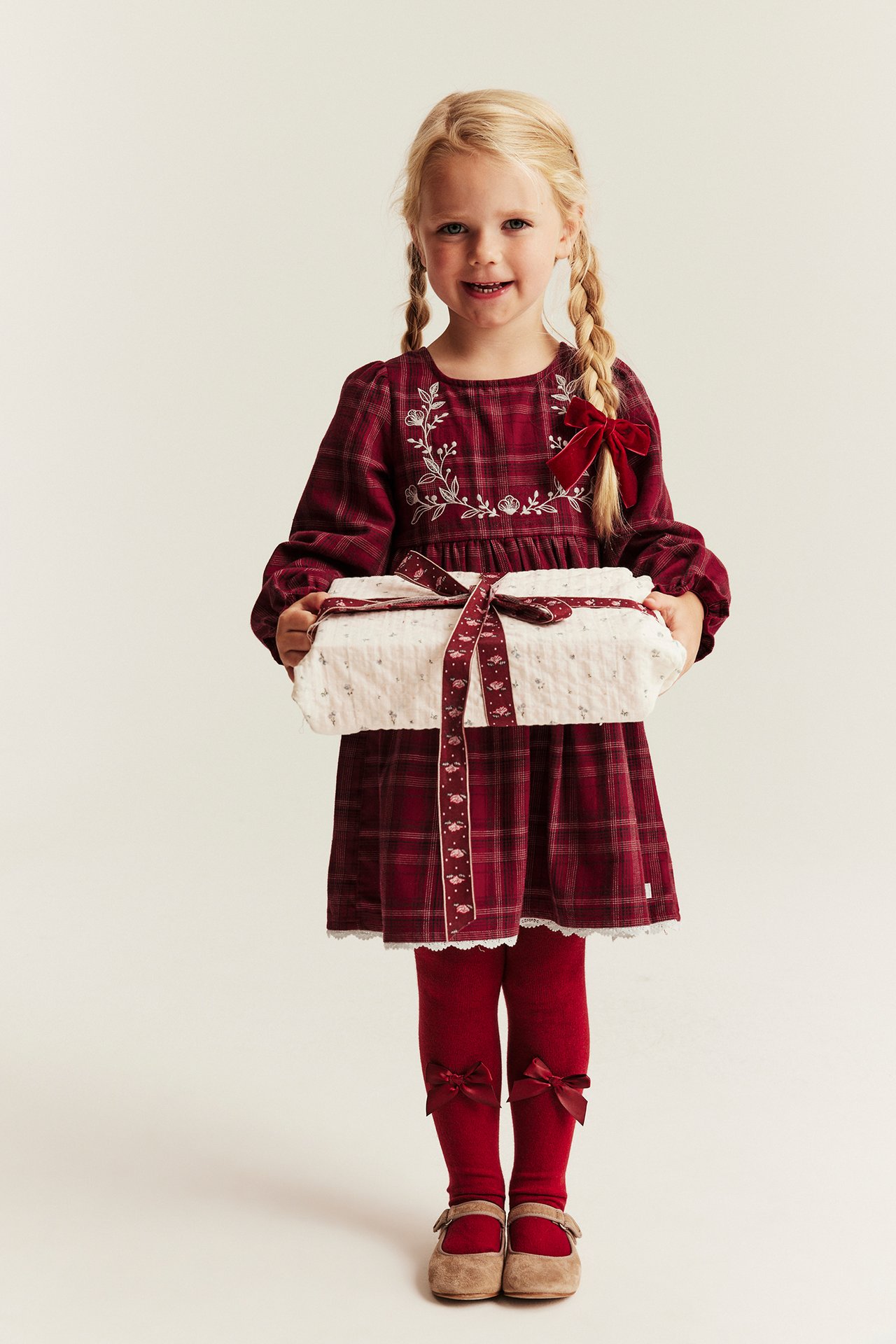 Kid's red plaid embroidered dress, red tights with bows, and beige shoes, holding a white present.