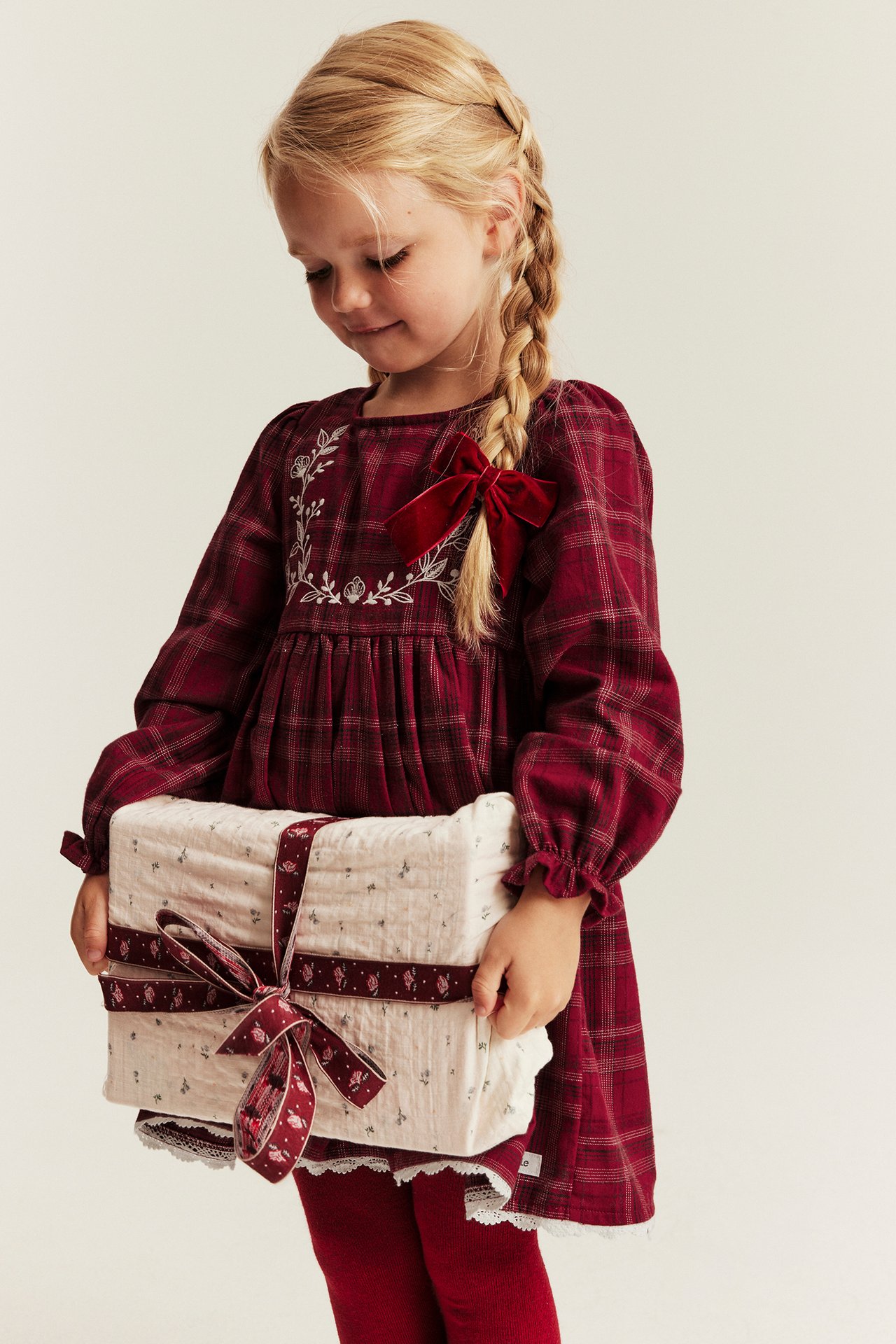 Front view of a girl in a red plaid dress with embroidery and velvet bow, holding a wrapped gift.