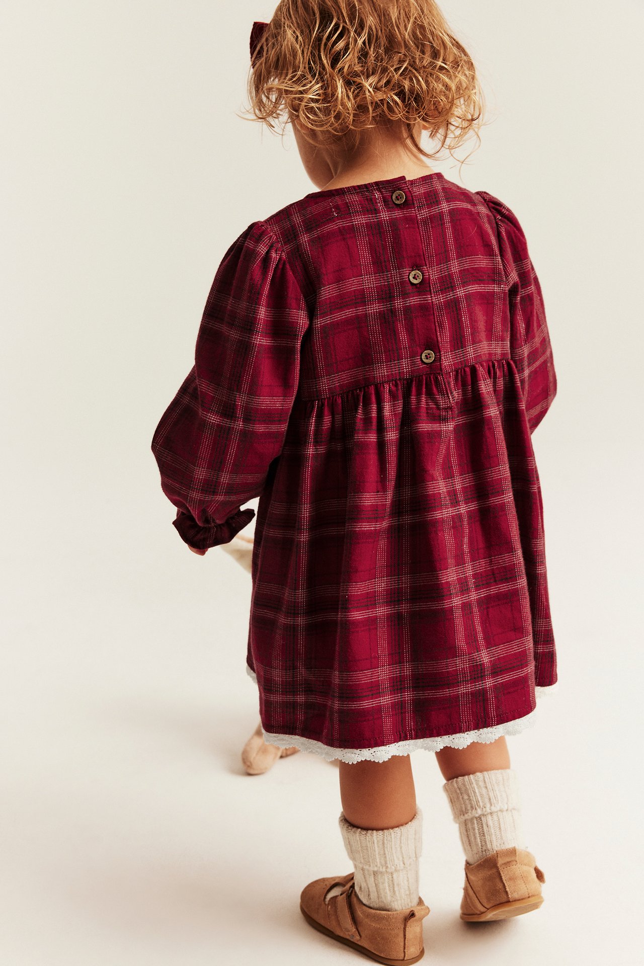 Rückansicht eines Kindes in rot-kariertem Langarmkleid mit weißer Spitze, Strickstulpen und braunen Schuhen.