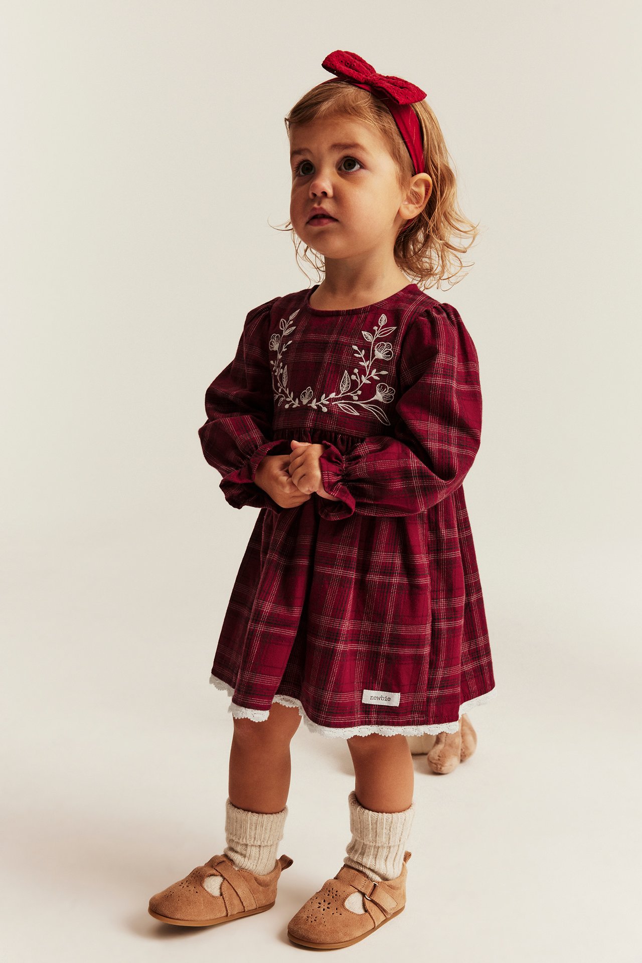 Vorderansicht: Baby-Mädchen in rot-kariertem besticktem Kleid, rotem Stirnband, beigen Socken, braunen Schuhen.
