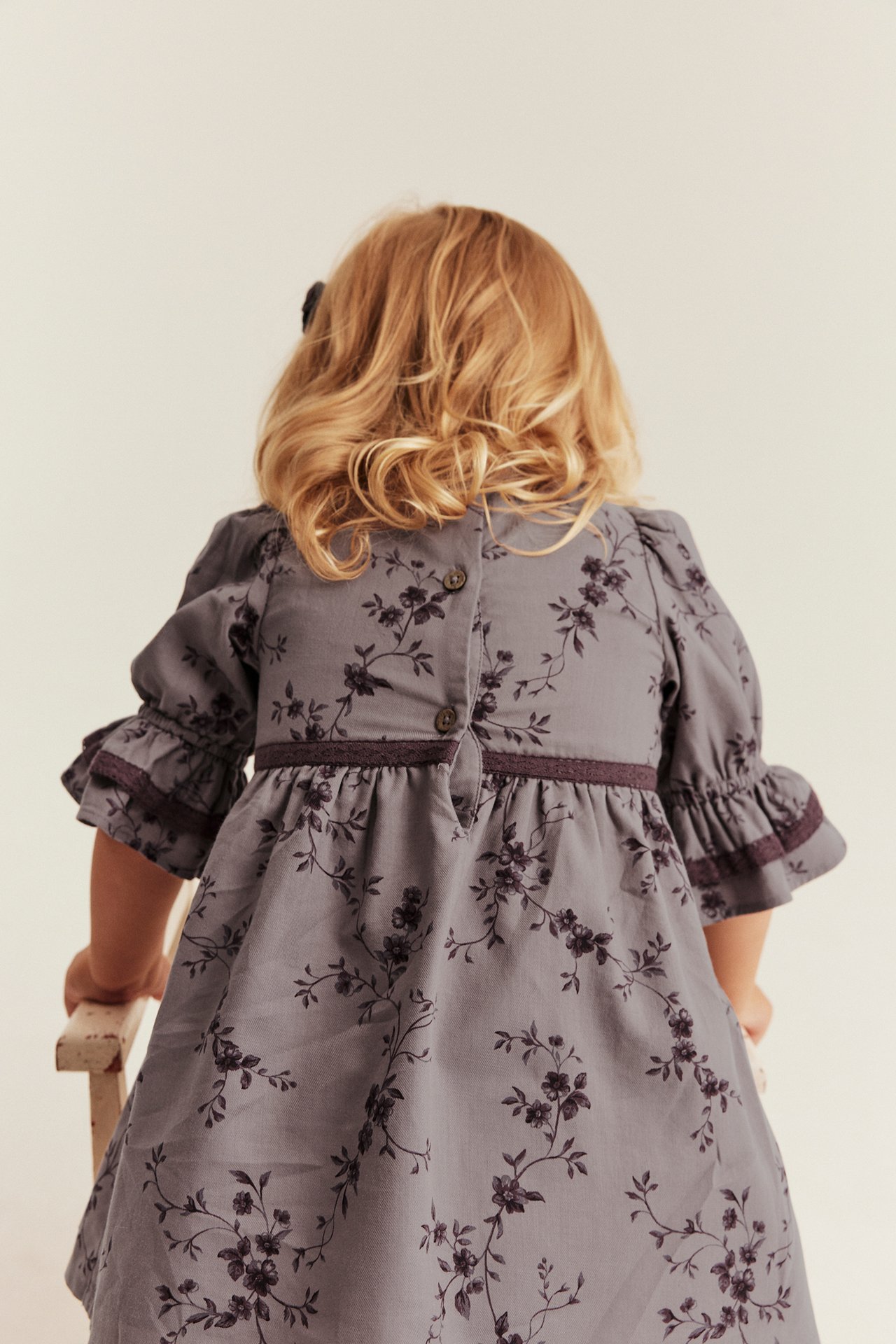 Rückansicht eines Kindes in grauem Blumenkleid mit Puffärmeln, Rüschenmanschetten und Knöpfen am Rücken.
