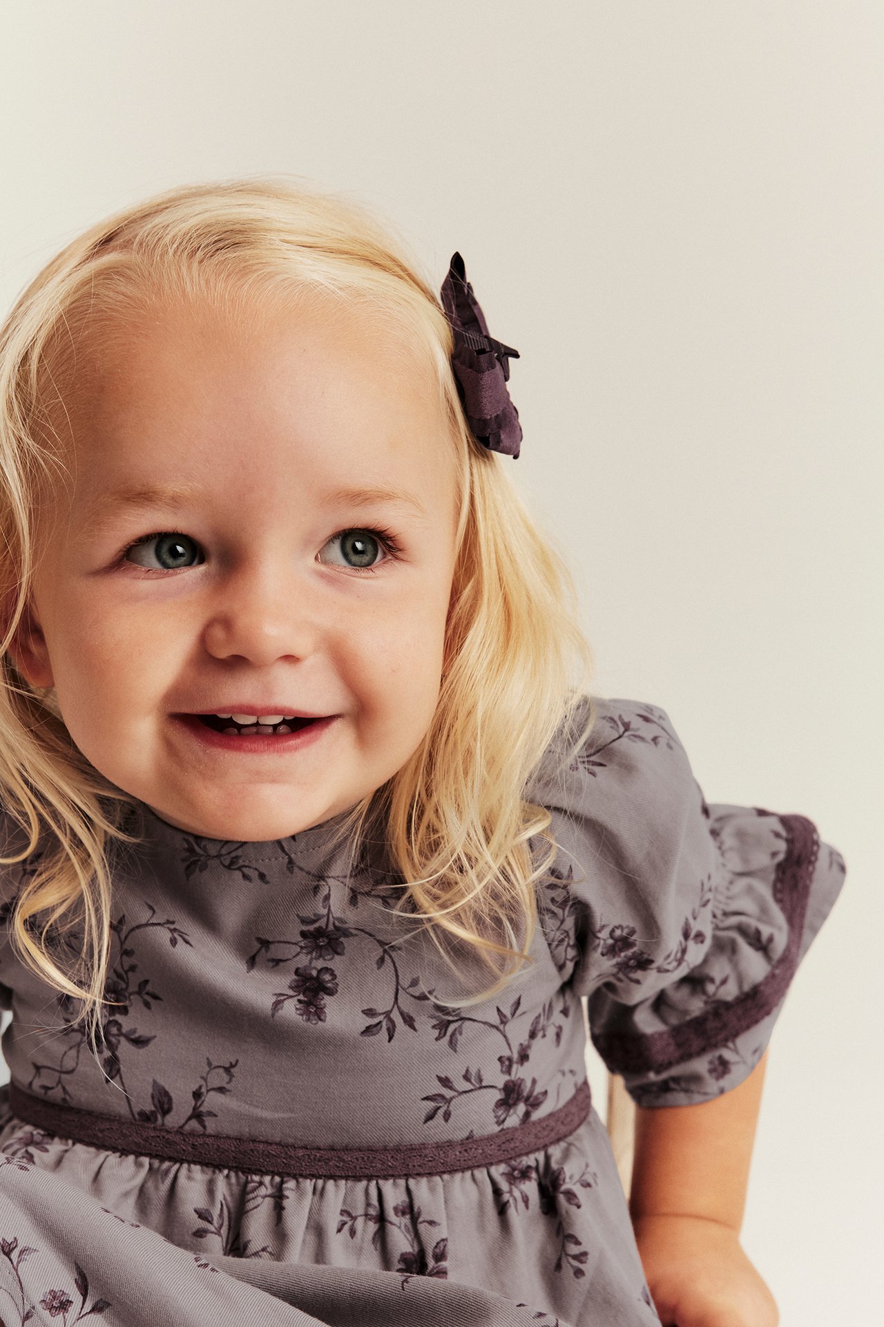 Nahaufnahme eines Kindes in einem mauve-grauen Blumenkleid mit Puffärmeln und dunklem Haarschmuck.