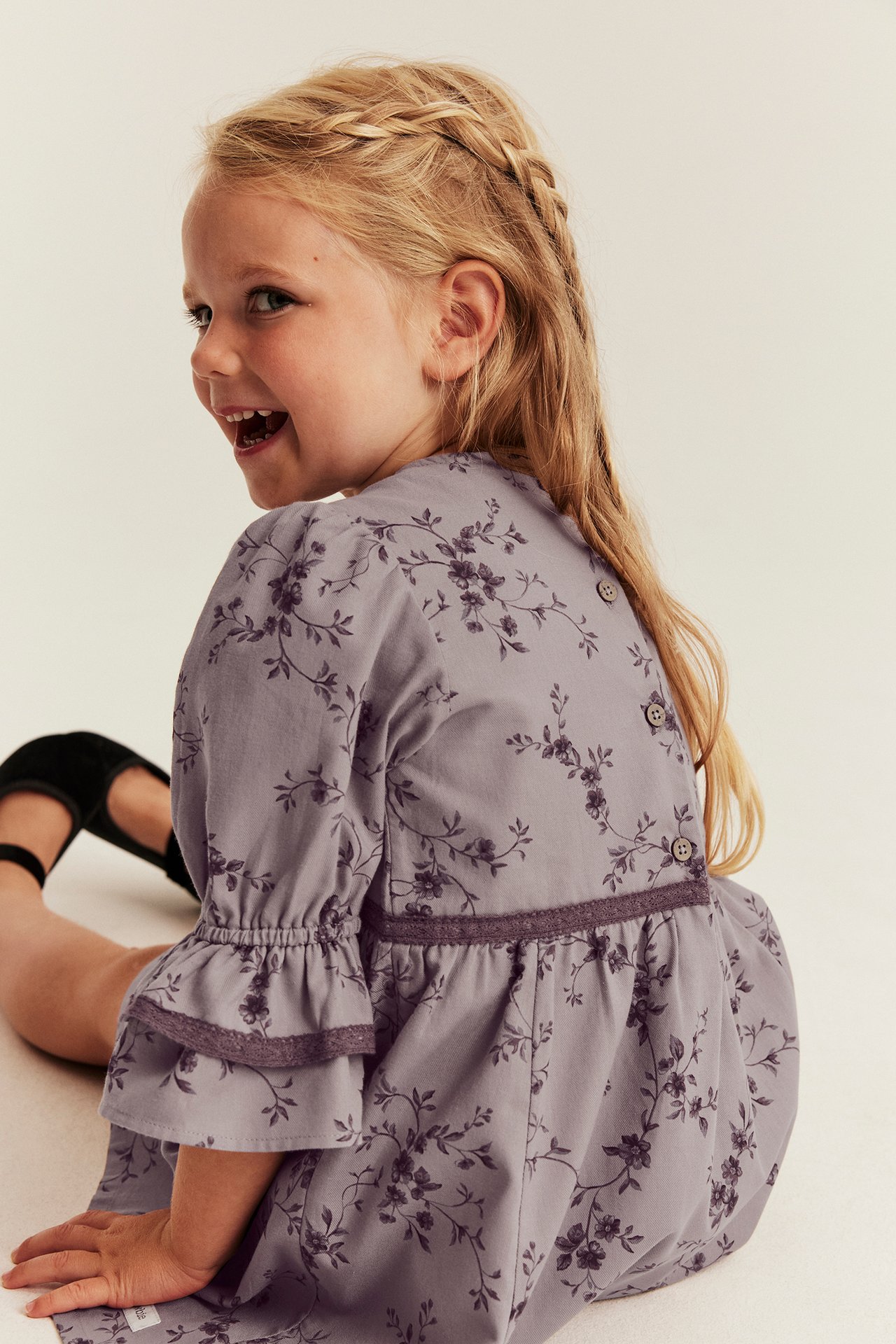 Back view of a kid wearing a purple floral dress with bell sleeves and button-down back, with black shoes.