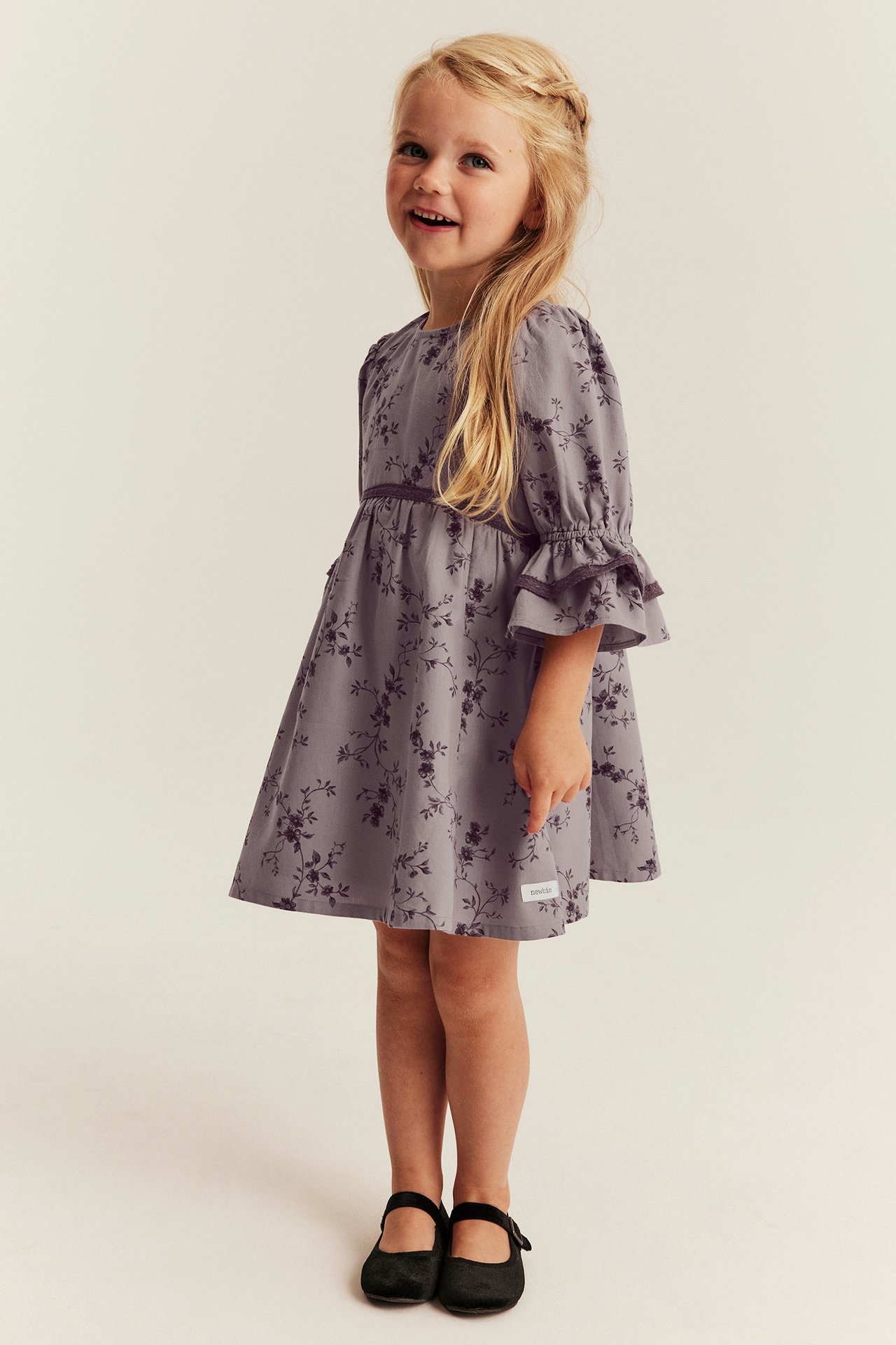 Full front view of a kid in a floral print purple dress with ruffled sleeves and black shoes.