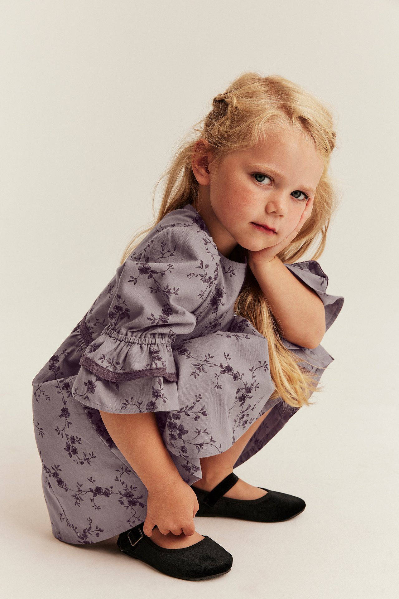 Front view of a kid wearing a lilac floral dress with ruffled sleeves and black flat shoes.