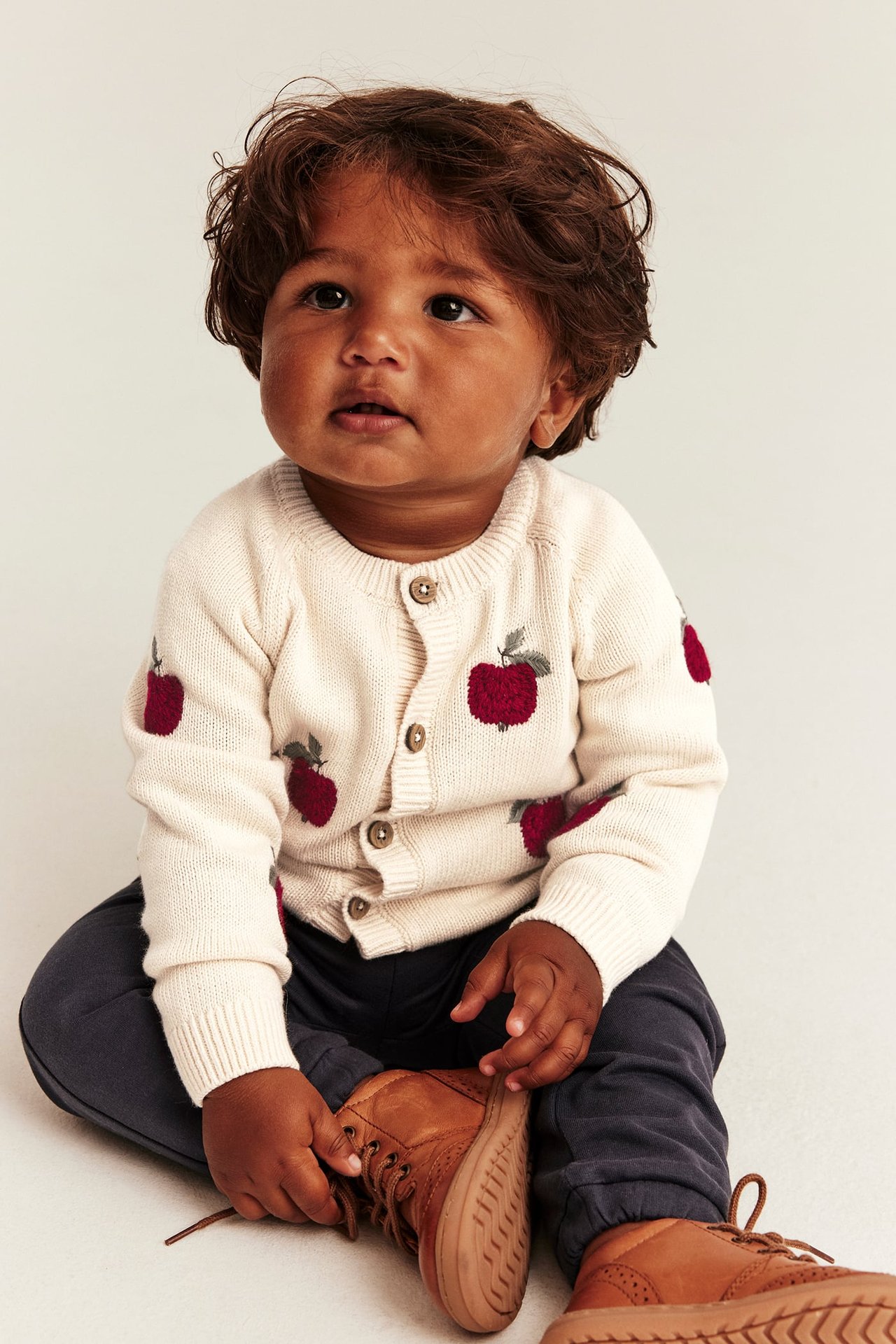 Front view of a baby wearing a cream knit cardigan with red apple print, dark pants, and brown lace-up shoes.
