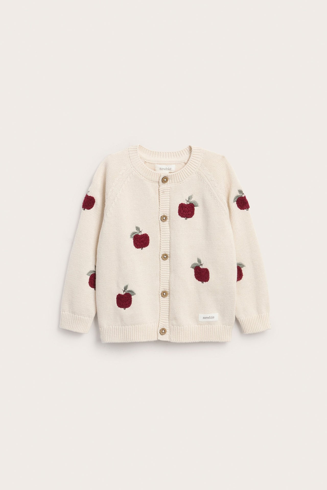 Vorderansicht einer cremefarbenen Baby-Strickjacke mit roter Apfelstickerei und braunen Knöpfen.