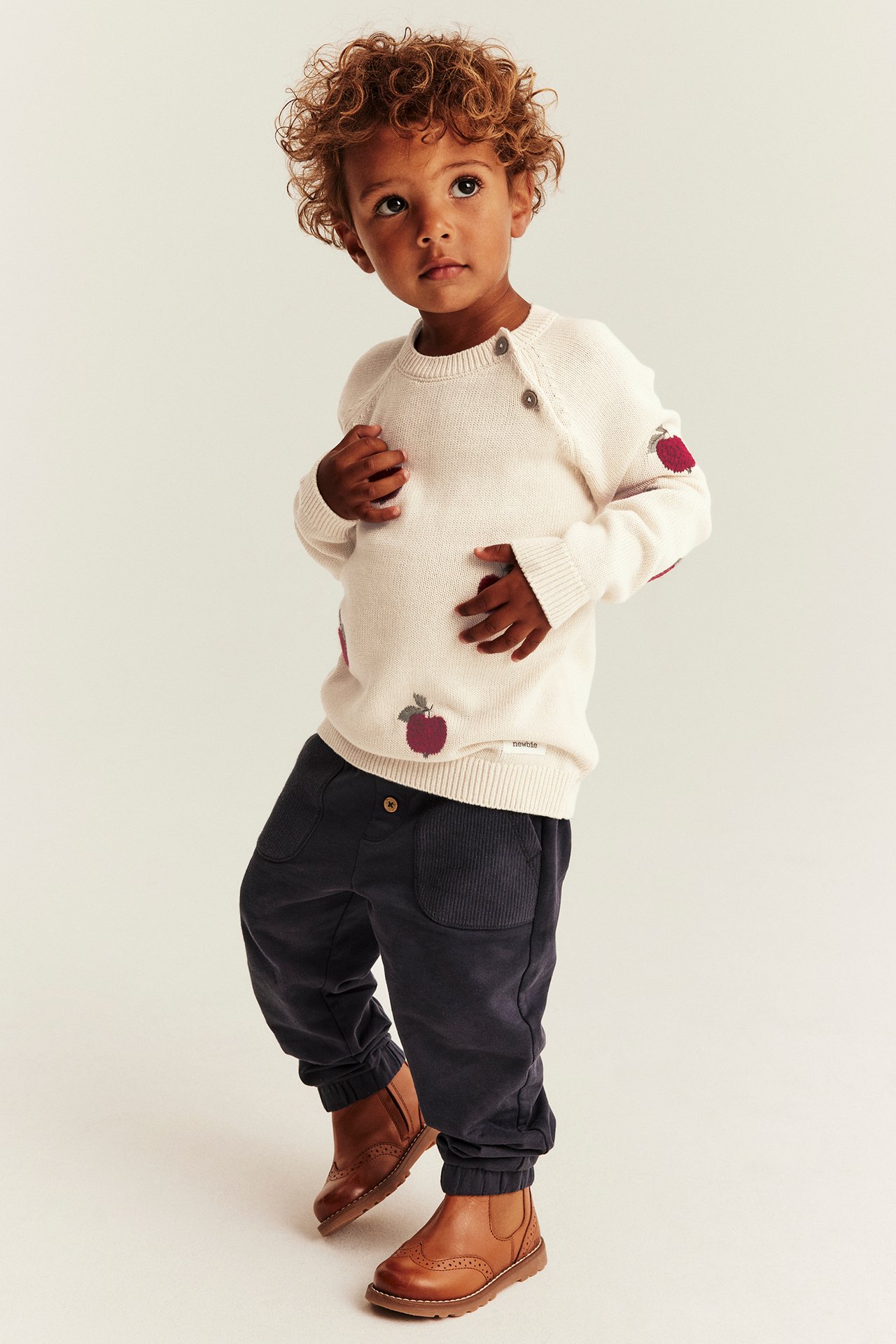 Frontansicht Kind in cremefarbenem Pullover, roten Apfelprints, dunkelblauer Cordhose und braunen Lederstiefeln.