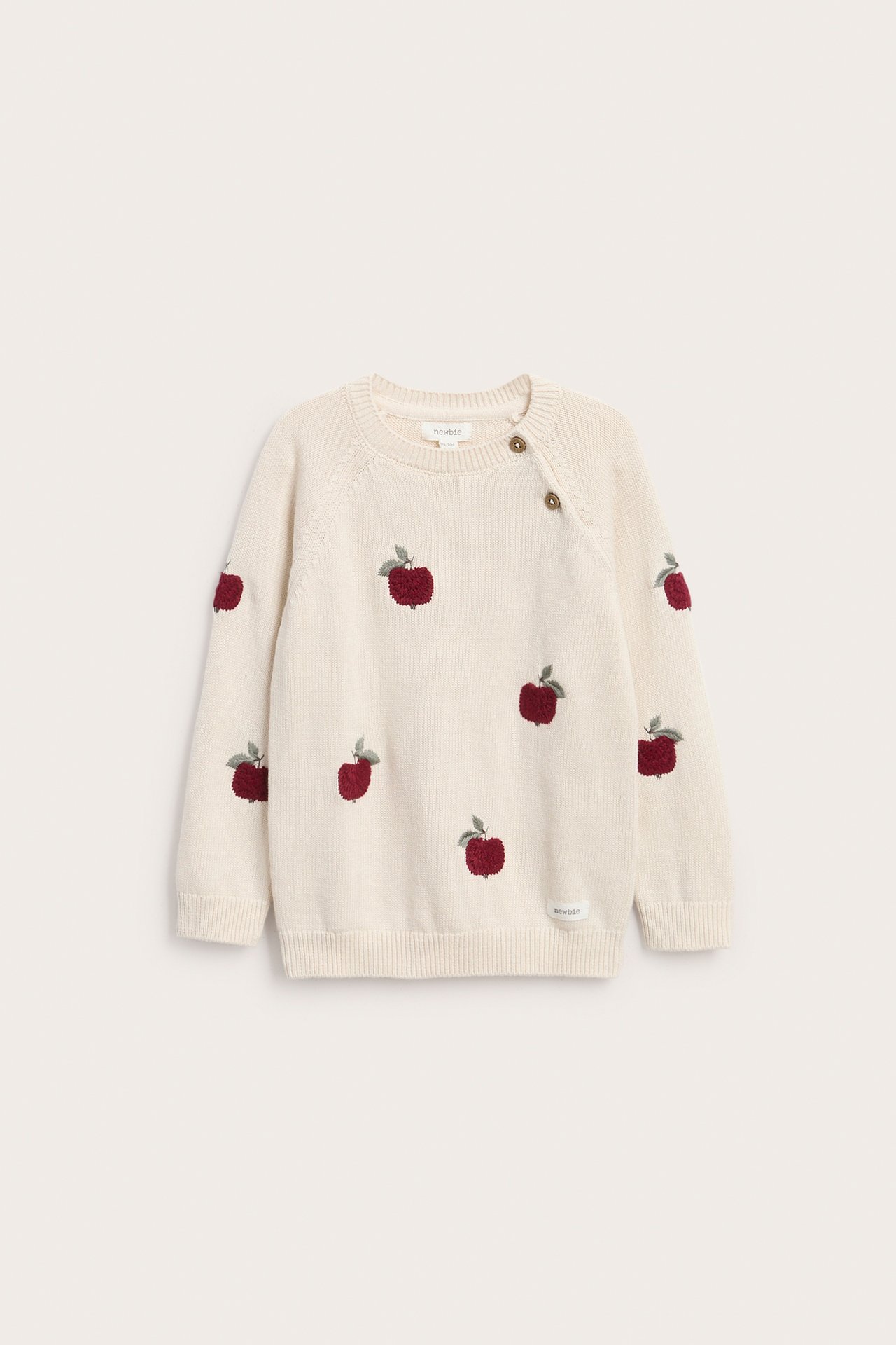 Vorderansicht eines cremefarbenen Baby-Pullover mit roter Apfelstickerei und Schulterknöpfen.