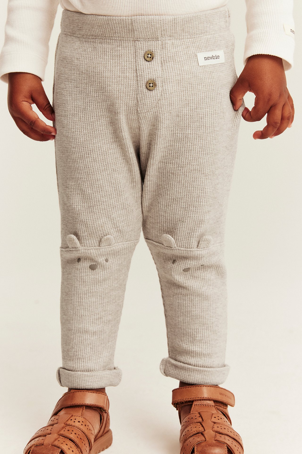 Kind in beige Waffel-Leggings mit Knie-Ohren, Frontknöpfen, einem weißen Shirt und braunen Sandalen.