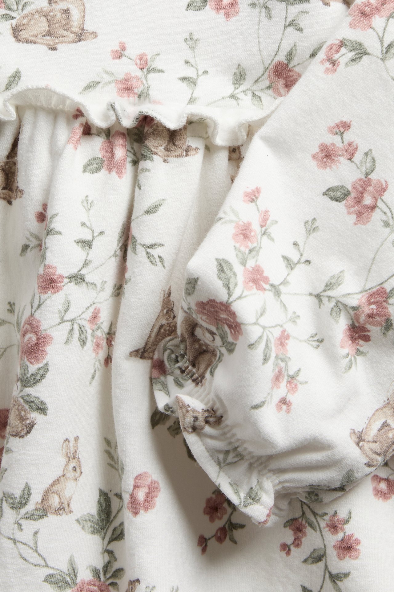 Close-up of an off-white kid's jersey dress with brown rabbit and pink floral pattern, featuring a ruffled edge.