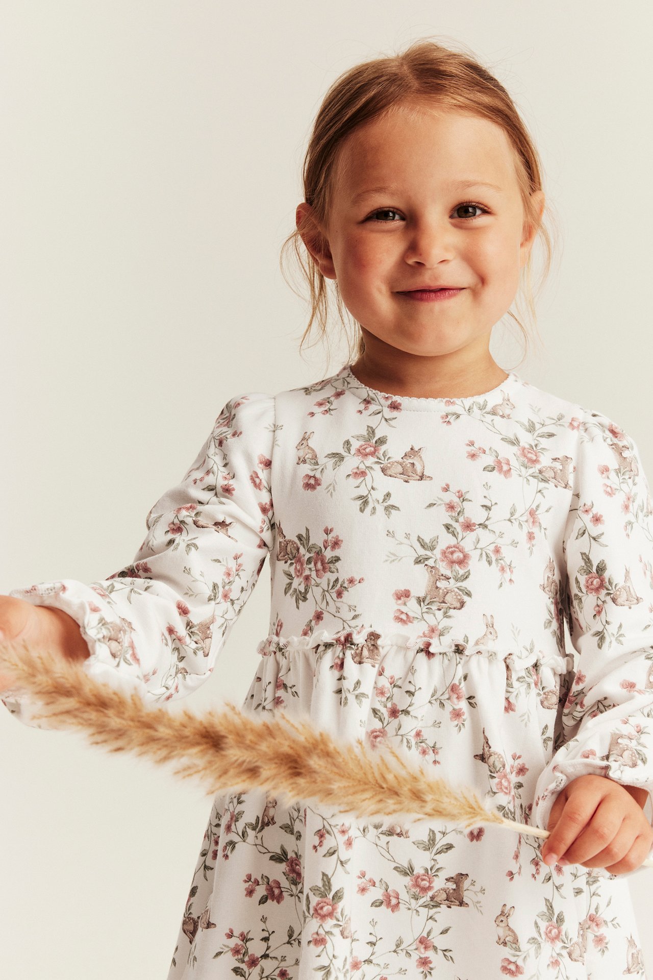 Front view of a kid wearing an off-white jersey dress with floral and rabbit pattern, holding a dried plant.