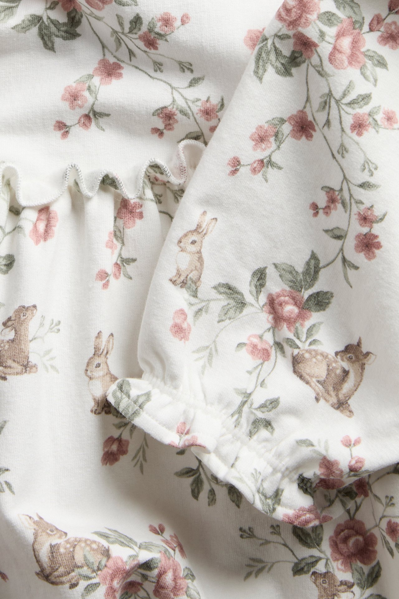 Close-up of an off-white baby jersey dress with pink floral, deer, and rabbit pattern, featuring ruffled cuffs.