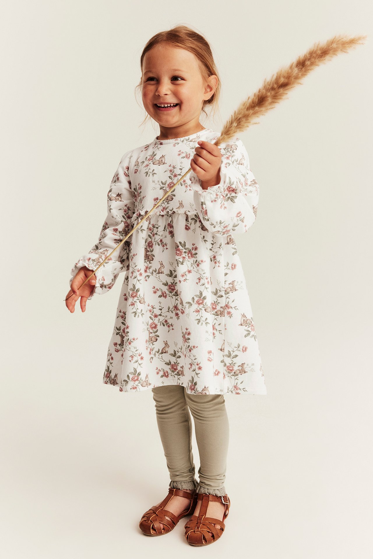 Kid in offwhite floral-deer jersey dress, light green leggings, brown sandals, holding a plant.