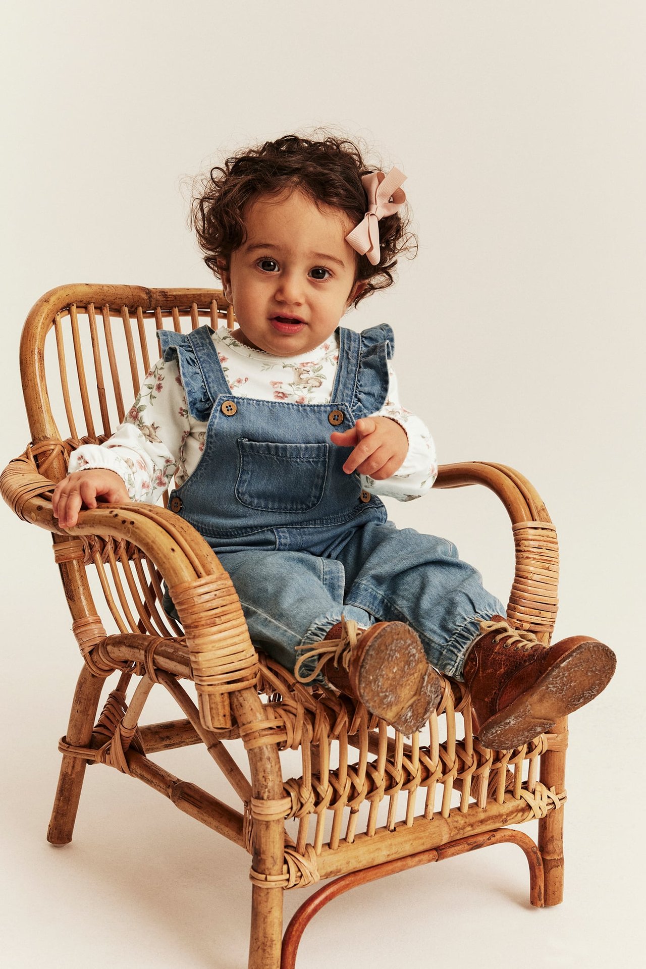 Front view baby in blue denim overalls, white floral top, brown lace-up boots, and pink bow.