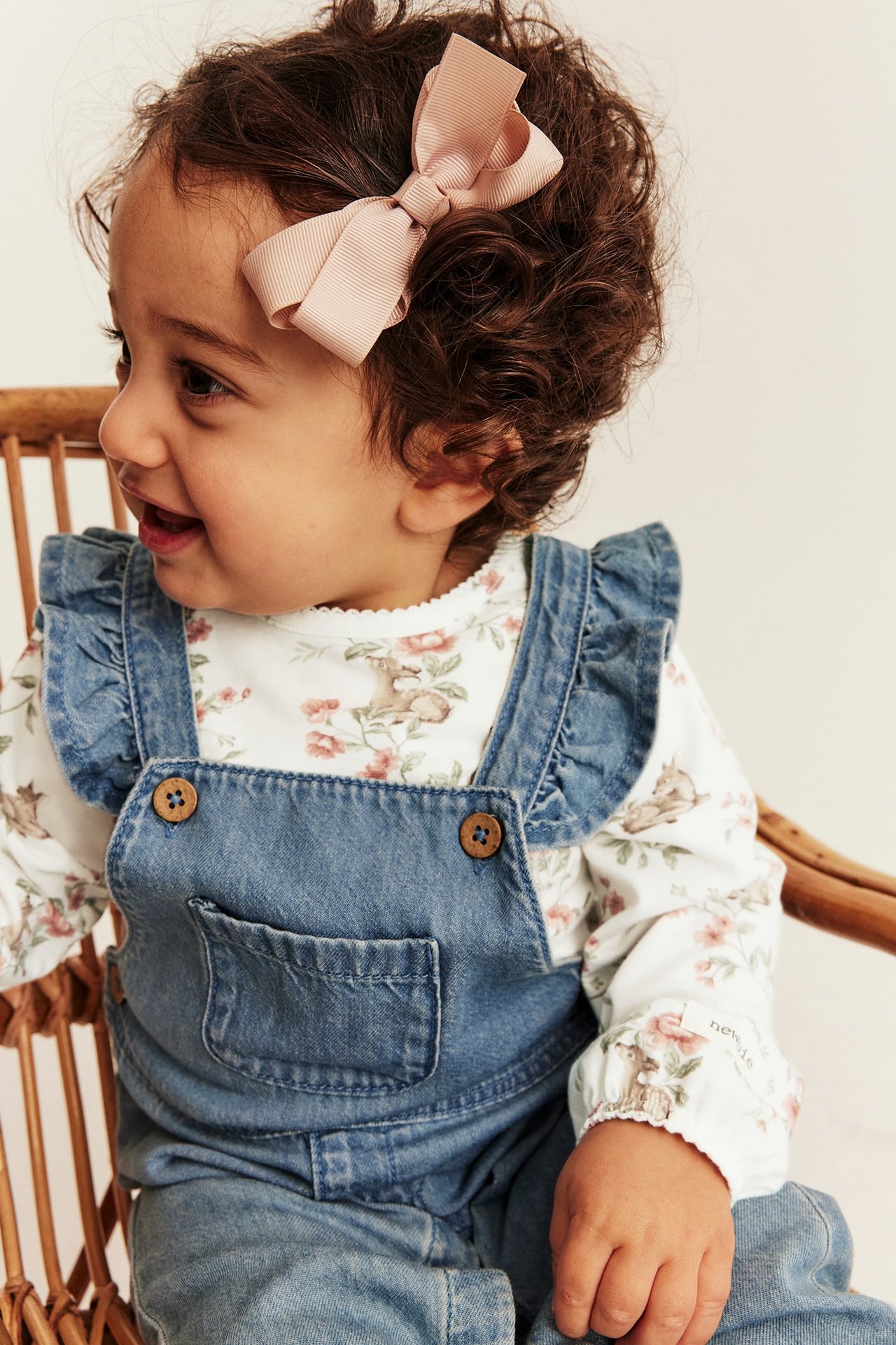 Close-up of baby wearing blue denim ruffled overalls, white floral long-sleeve top, and pink bow.