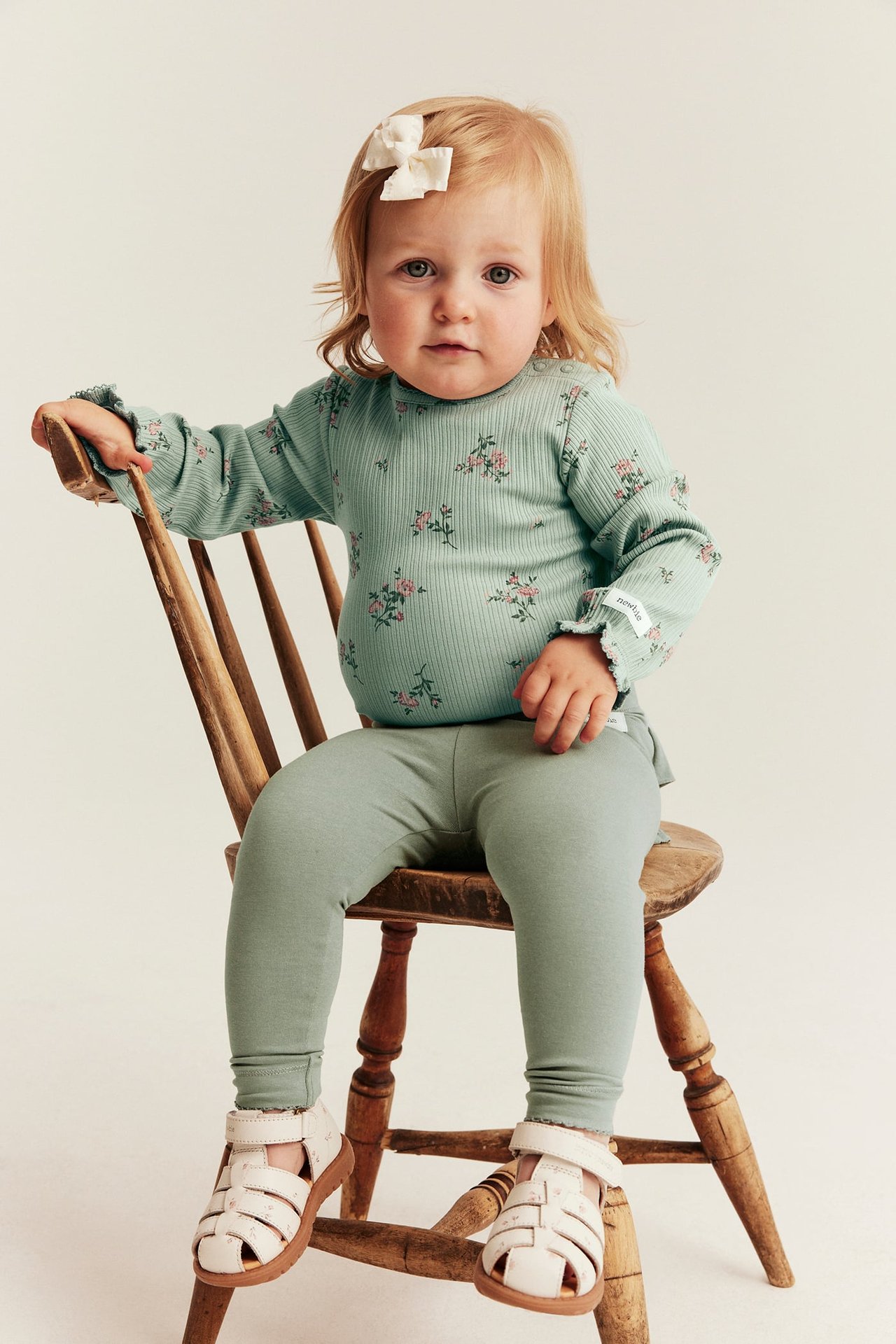 Front view baby in green floral ribbed long-sleeve top, green leggings, white sandals, white bow.