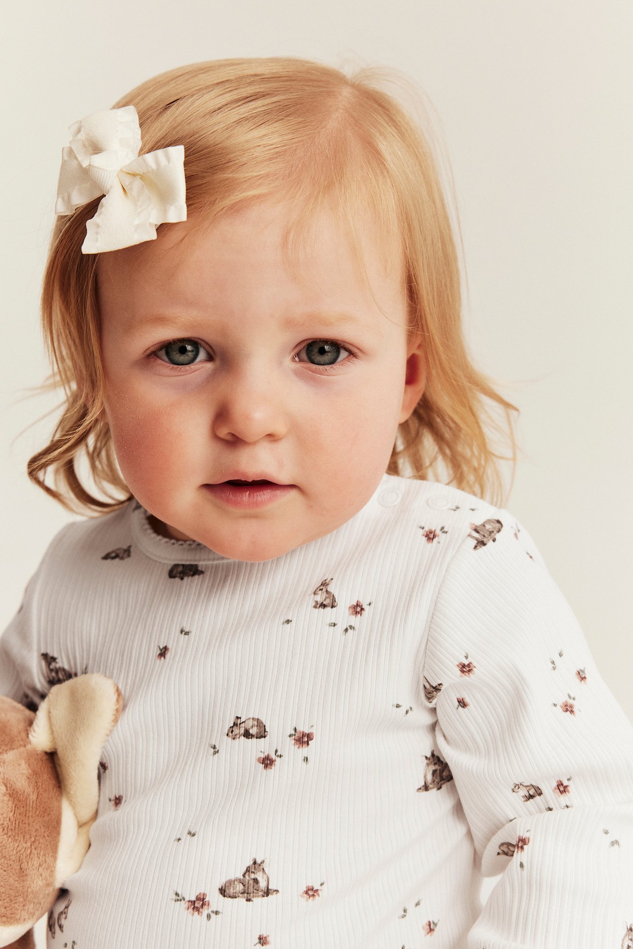 Front view of a baby in an offwhite ribbed body with animal/floral print, and an offwhite hair bow.