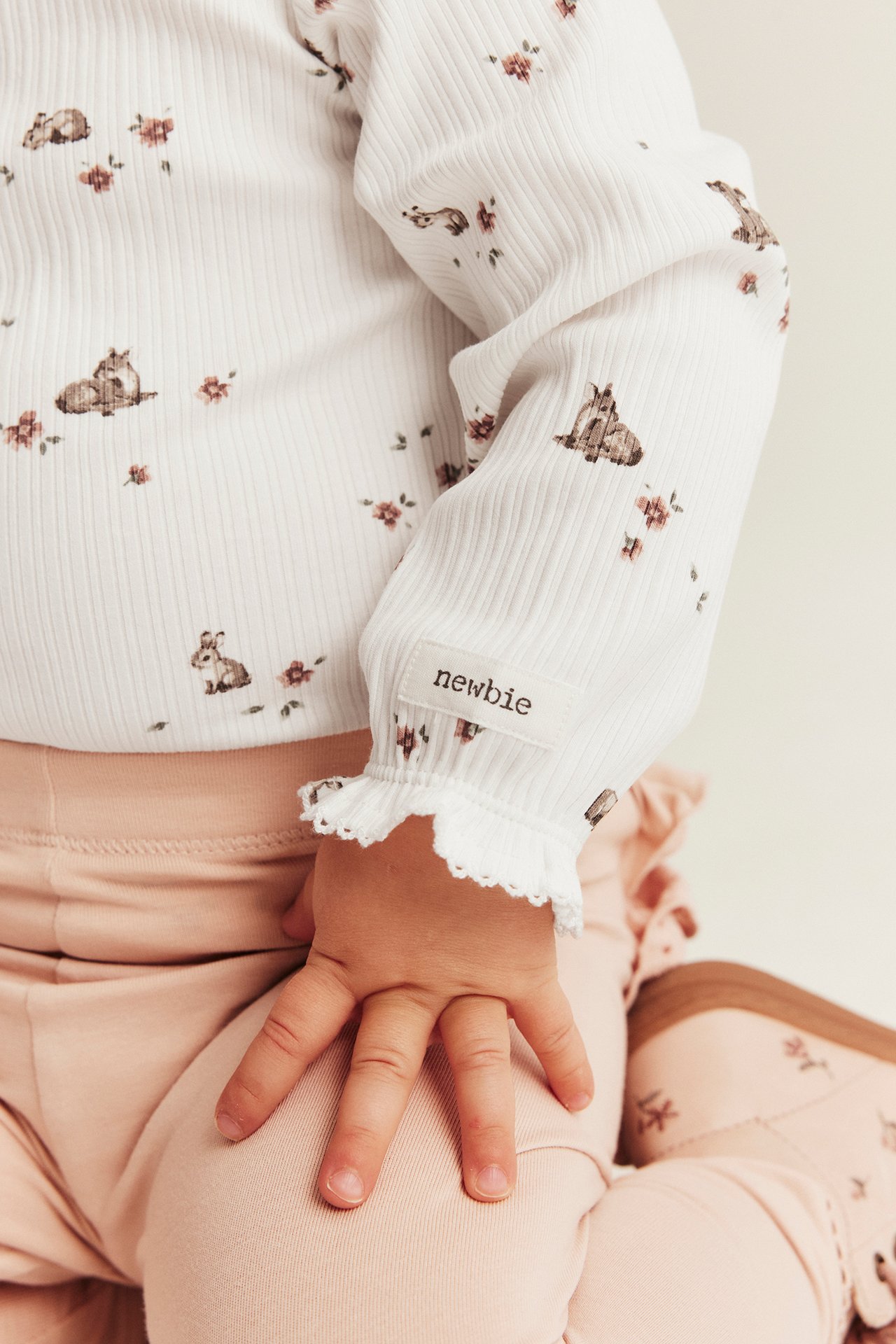 Close-up of baby in an off-white ribbed bodysuit with deer, floral print, ruffle cuffs, and pink trousers.