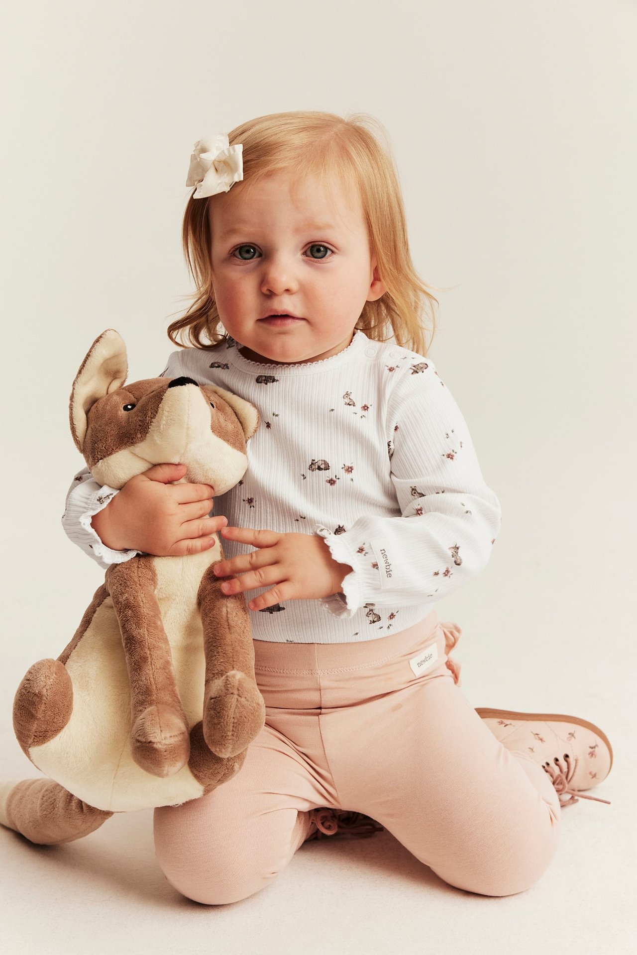 Front view, baby in off-white ribbed floral deer print bodysuit, light pink leggings, holding stuffed animal.