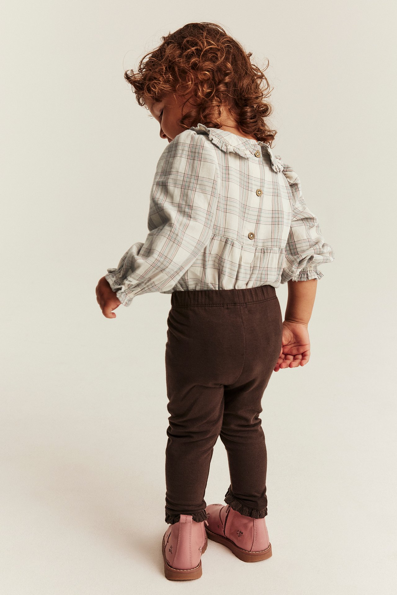 Back view of a kid wearing a plaid ruffled top, brown ruffled leggings, and pink shoes.