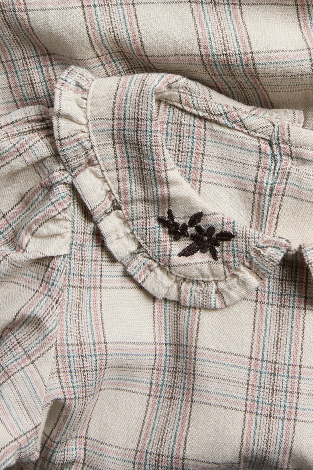 Close-up of a beige plaid top with a ruffled collar and dark brown floral embroidery.